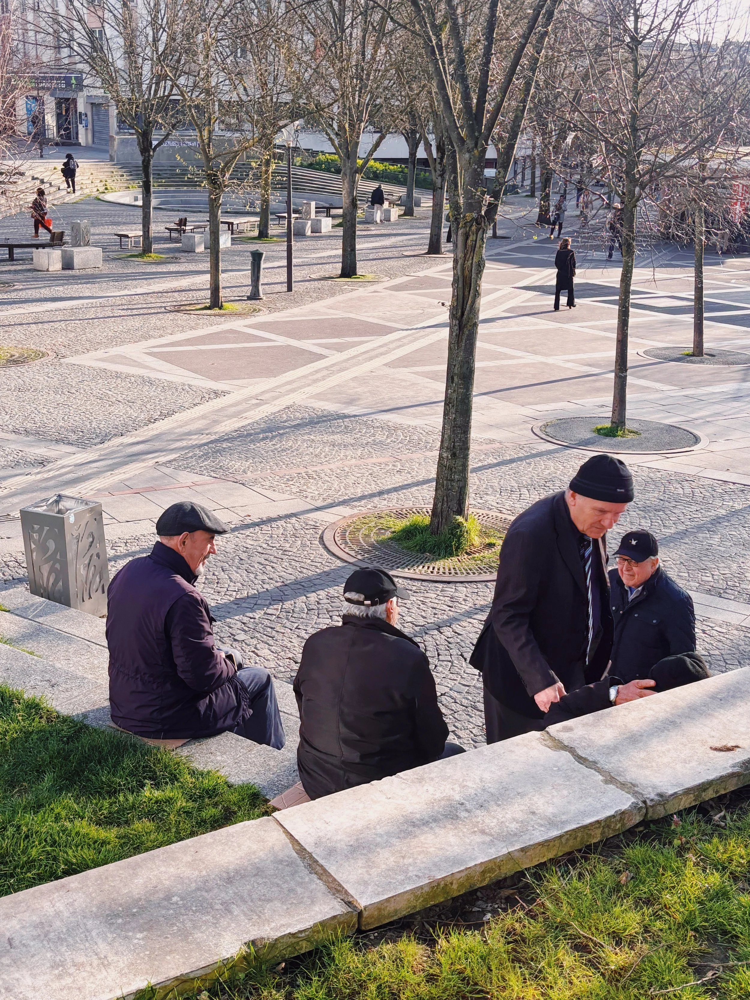 Groupe de personnes assises et conversant dans un parc urbain avec des arbres dénudés, quelques bancs et des piétons en arrière-plan.