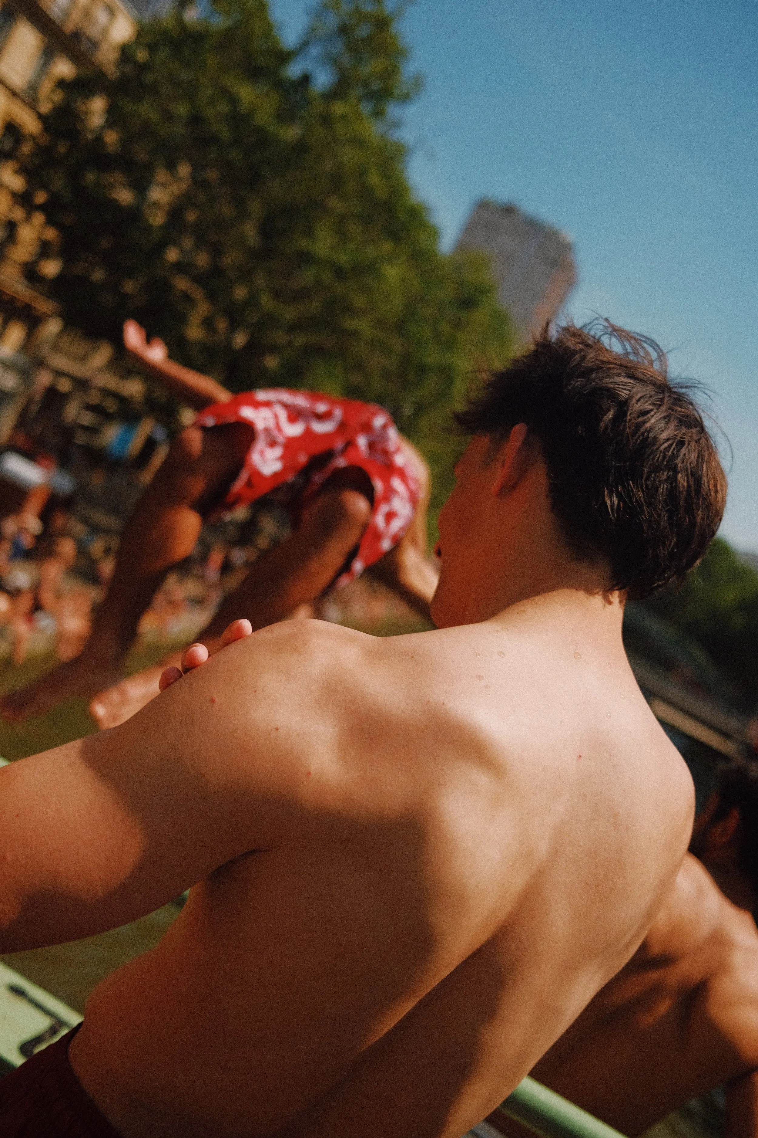 Jeune homme nu vu de dos, cheveux bruns, tenant un vêtement rouge à motifs blancs, en extérieur, avec un site touristique en arrière-plan et un arbre large.
