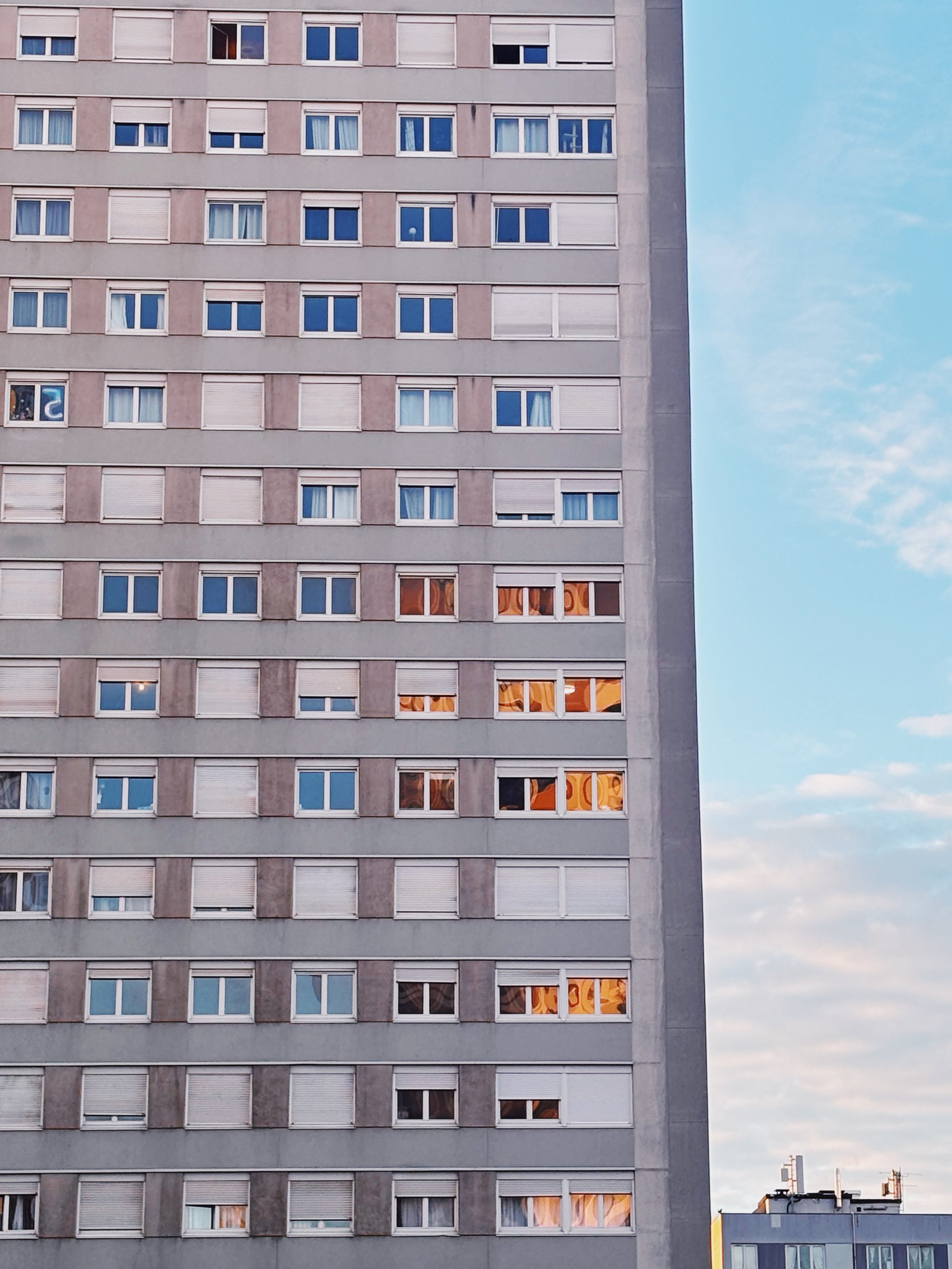 Immeuble résidentiel en face avec plusieurs fenêtres, certains volets sont fermés, un ciel bleu avec quelques nuages, et deux autres bâtiments plus petits en bas à droite.