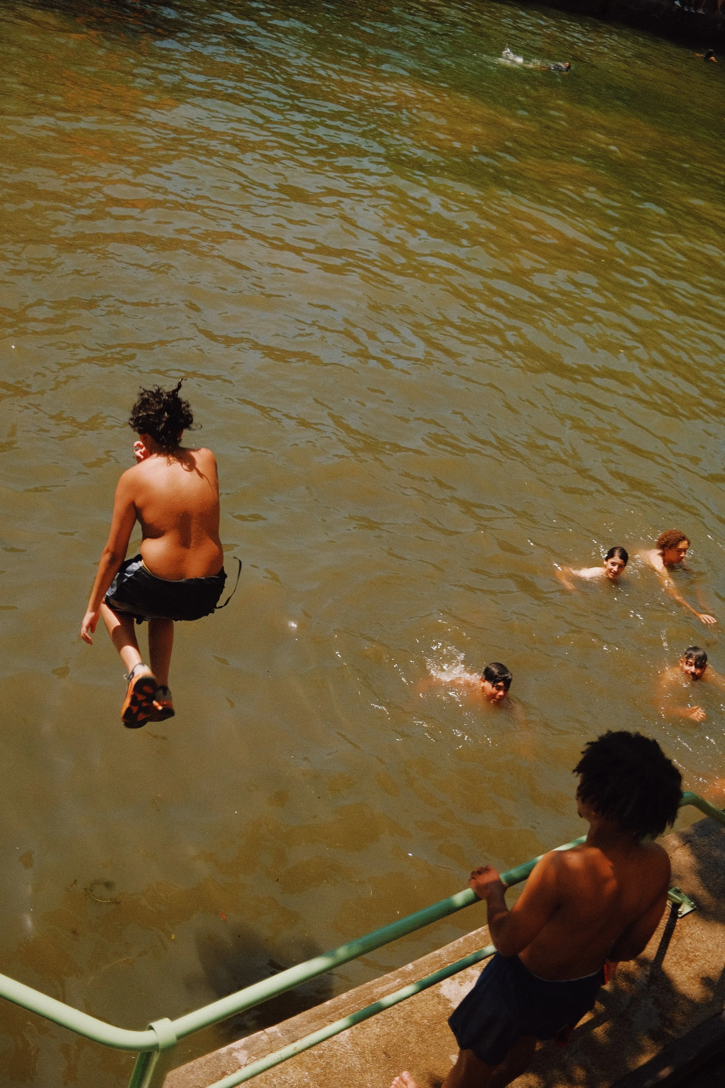 Groupe d’enfants nageant dans une rivière ou un lac, certains en train de nager, un garçon assis sur la rive avec une canne en PVC, et un autre qui saute dans l’eau.