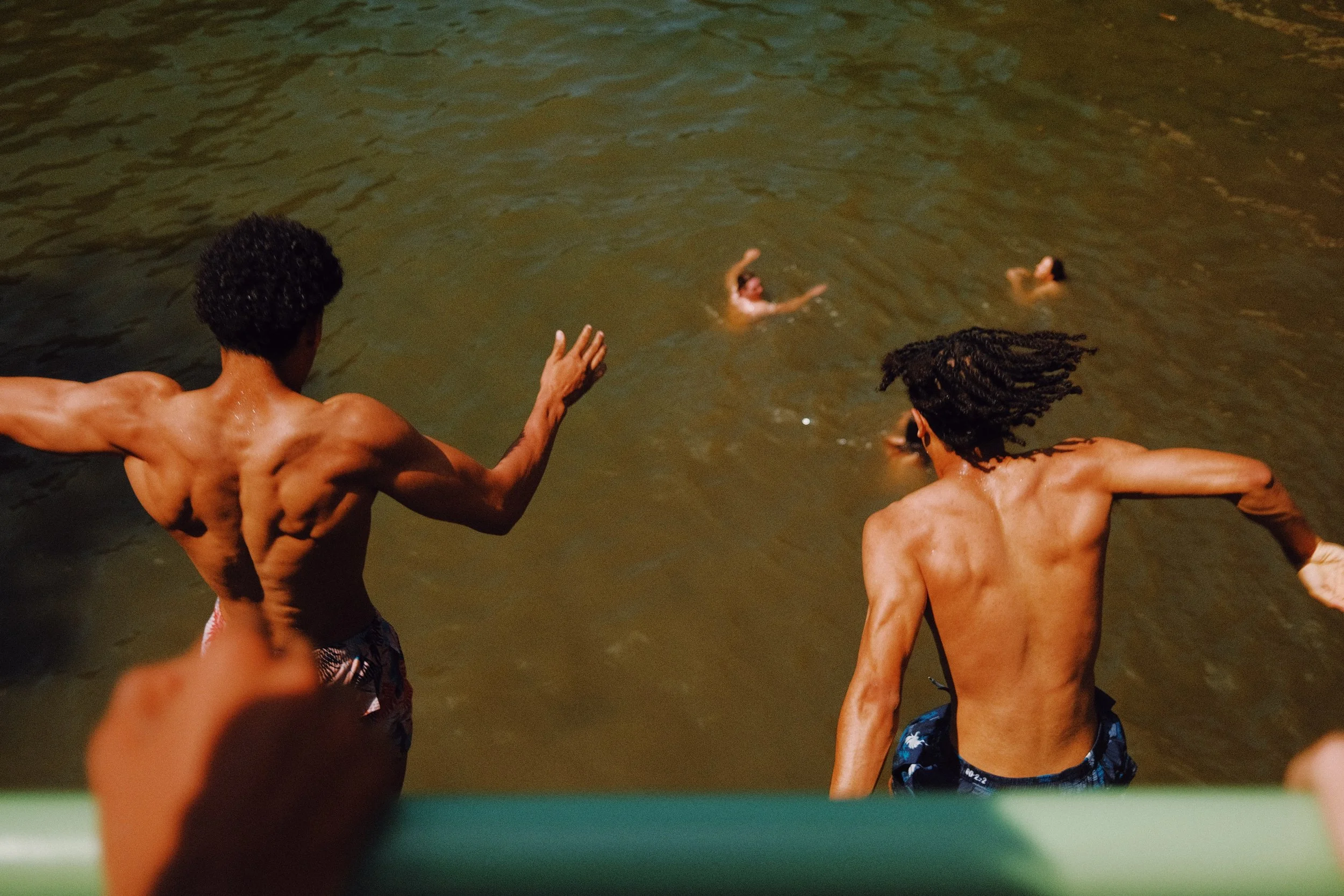 Deux hommes nus jouent dans l'eau d'une rivière ou d'un lac, avec une personne à l'arrière-plan nageant ou flottant.
