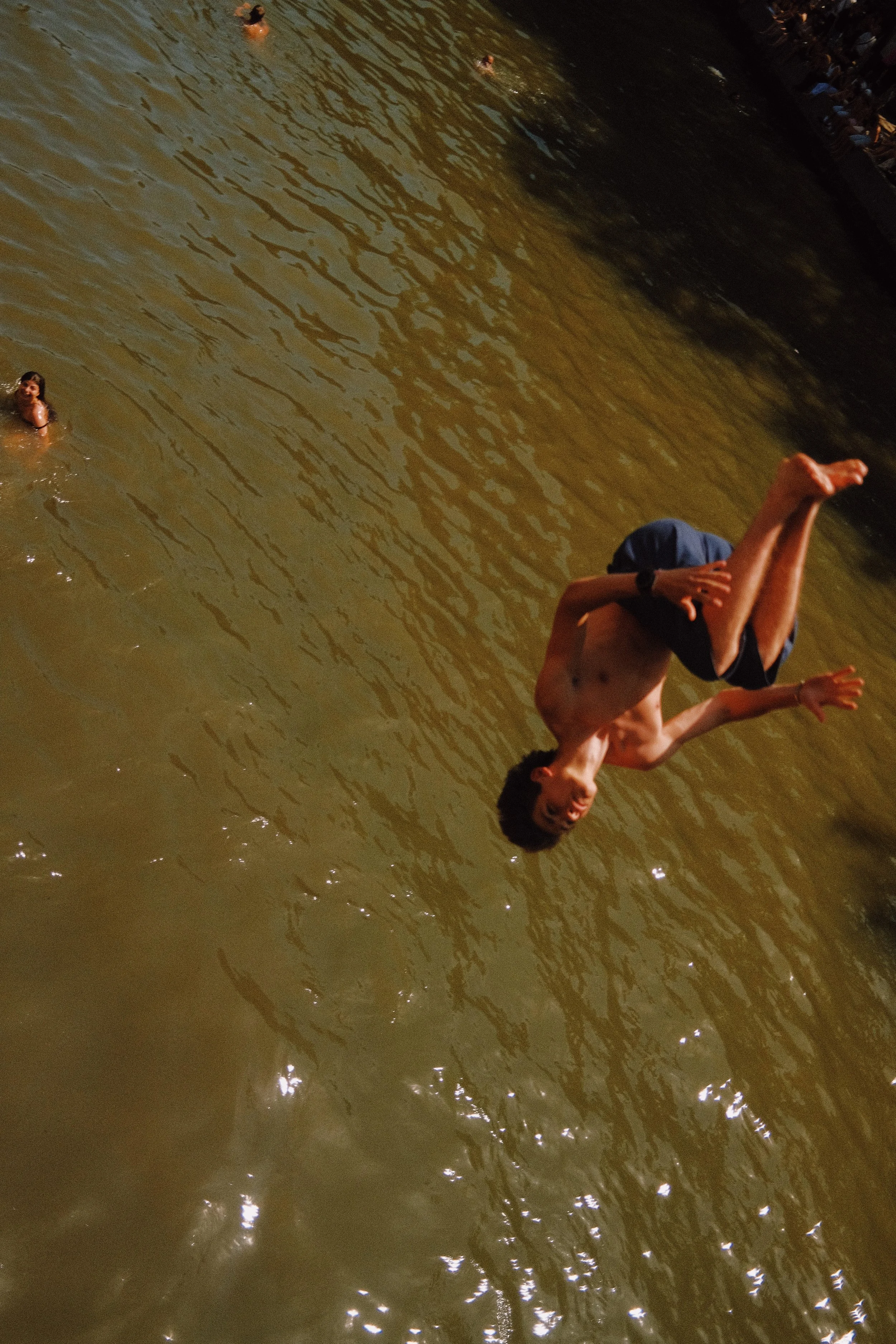 Personnage qui fait une acrobatie dans l'eau, d'autres personnes nagent à proximité dans une rivière, au coucher du soleil.