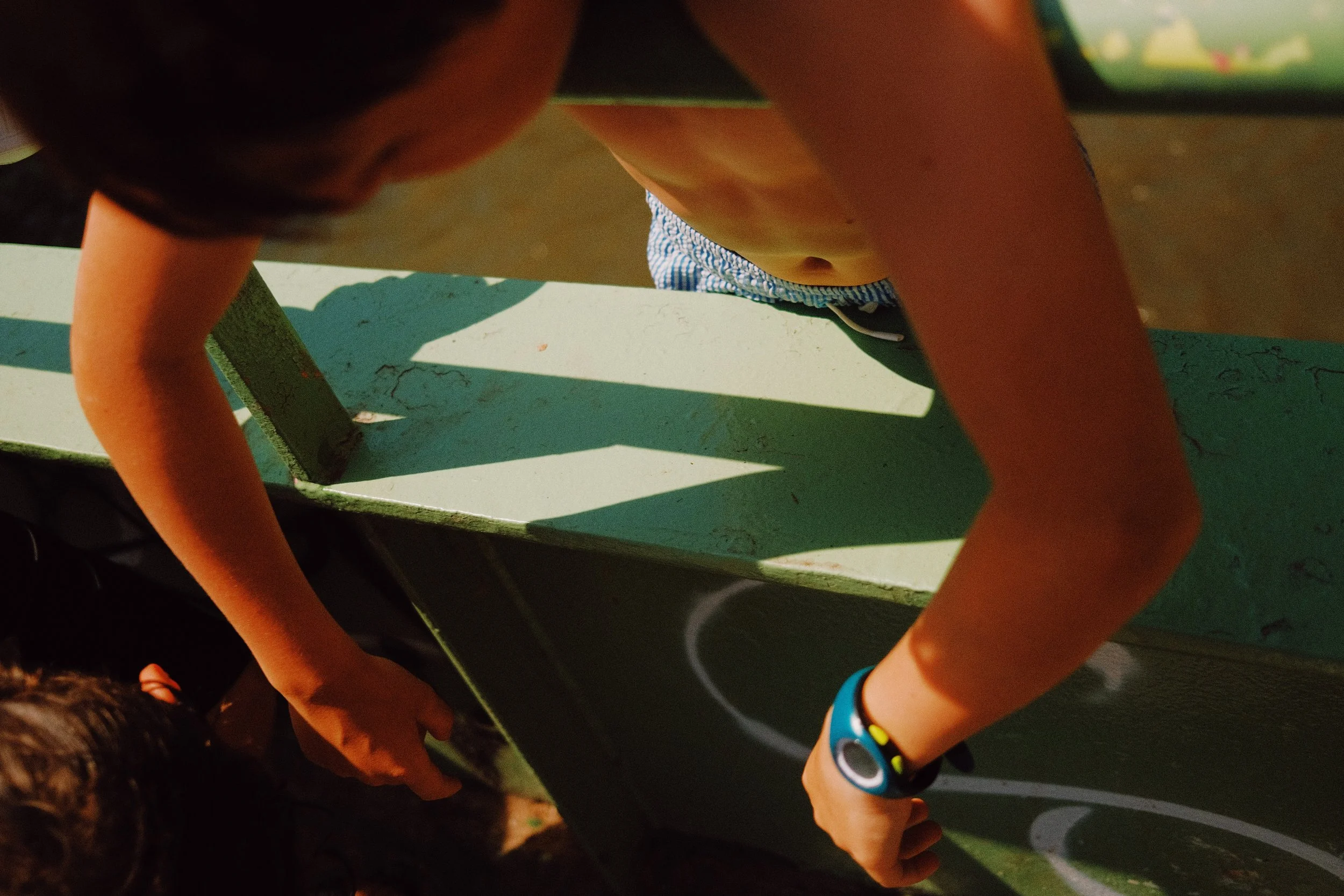 Les mains de plusieurs enfants qui se tiennent autour d'une table de pique-nique ou d'une surface de jeu, ensoleillée, avec des bracelets colorés.