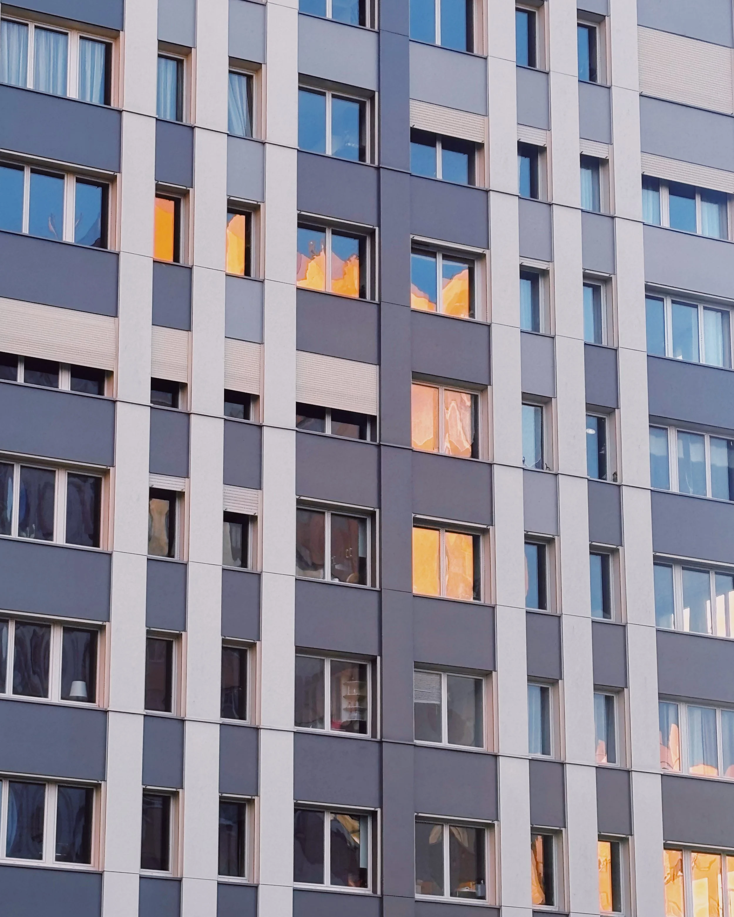 Façade d'un immeuble résidentiel avec plusieurs fenêtres, certaines reflètent un ciel orangeâtre, indiquant un coucher ou un lever de soleil.