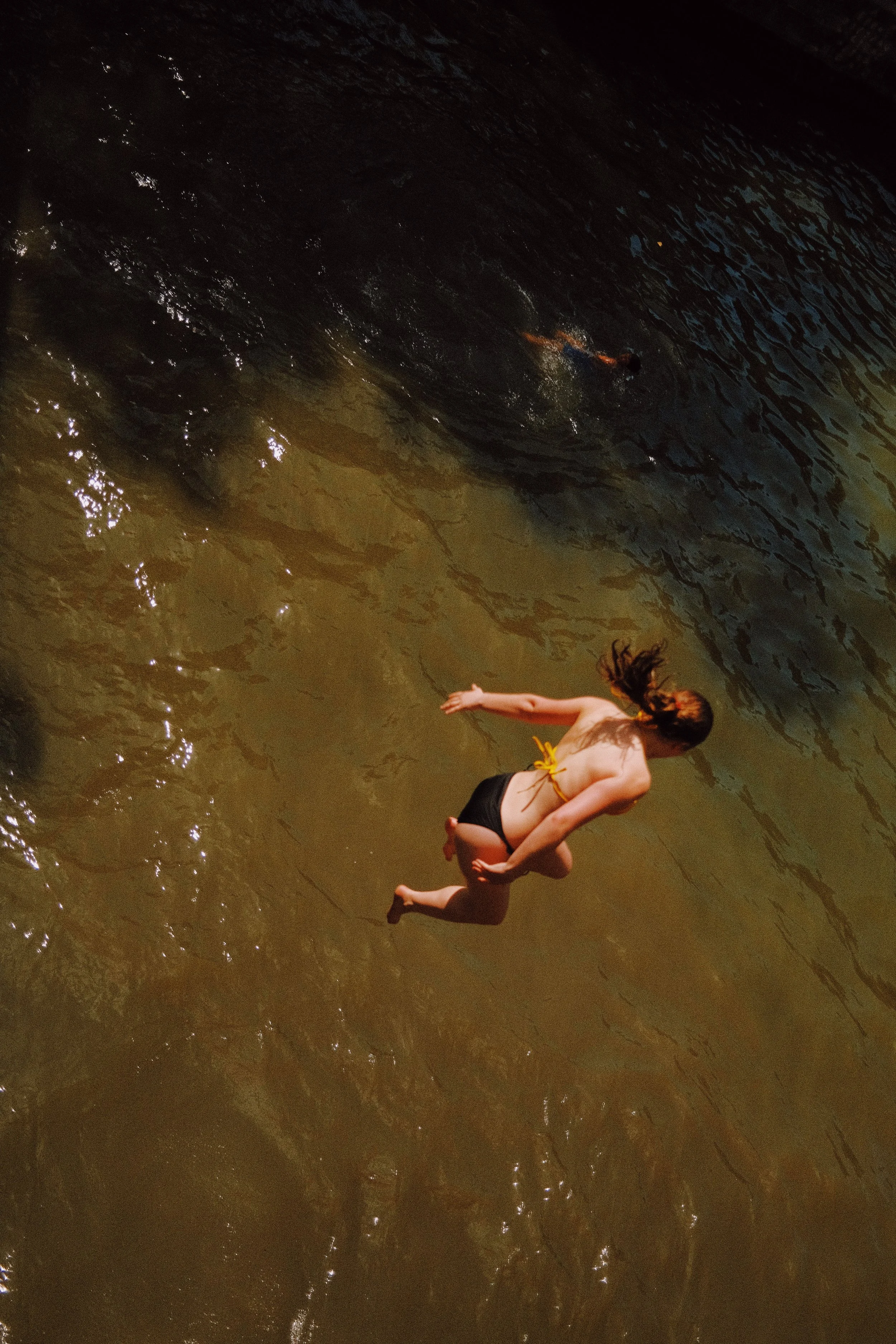 Une femme nue portant un maillot de bain jaune nage dans une rivière ou un lac avec de l'eau sombre et des rayons de lumière. La photo est prise en plongée.