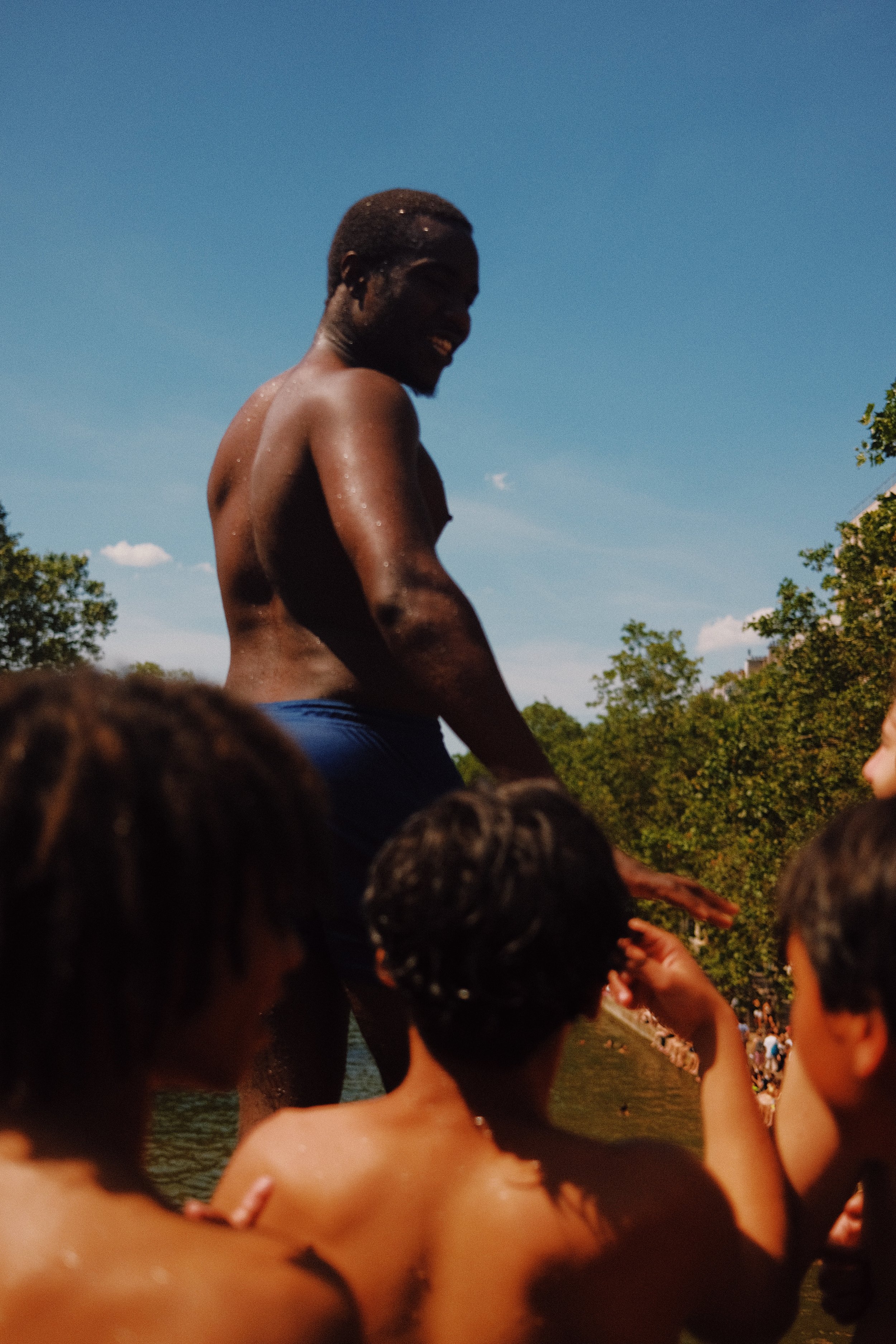 Un homme souriant, de peau foncée, est debout dans un groupe de personnes nues près de l'eau, avec un ciel clair et des arbres en arrière-plan.