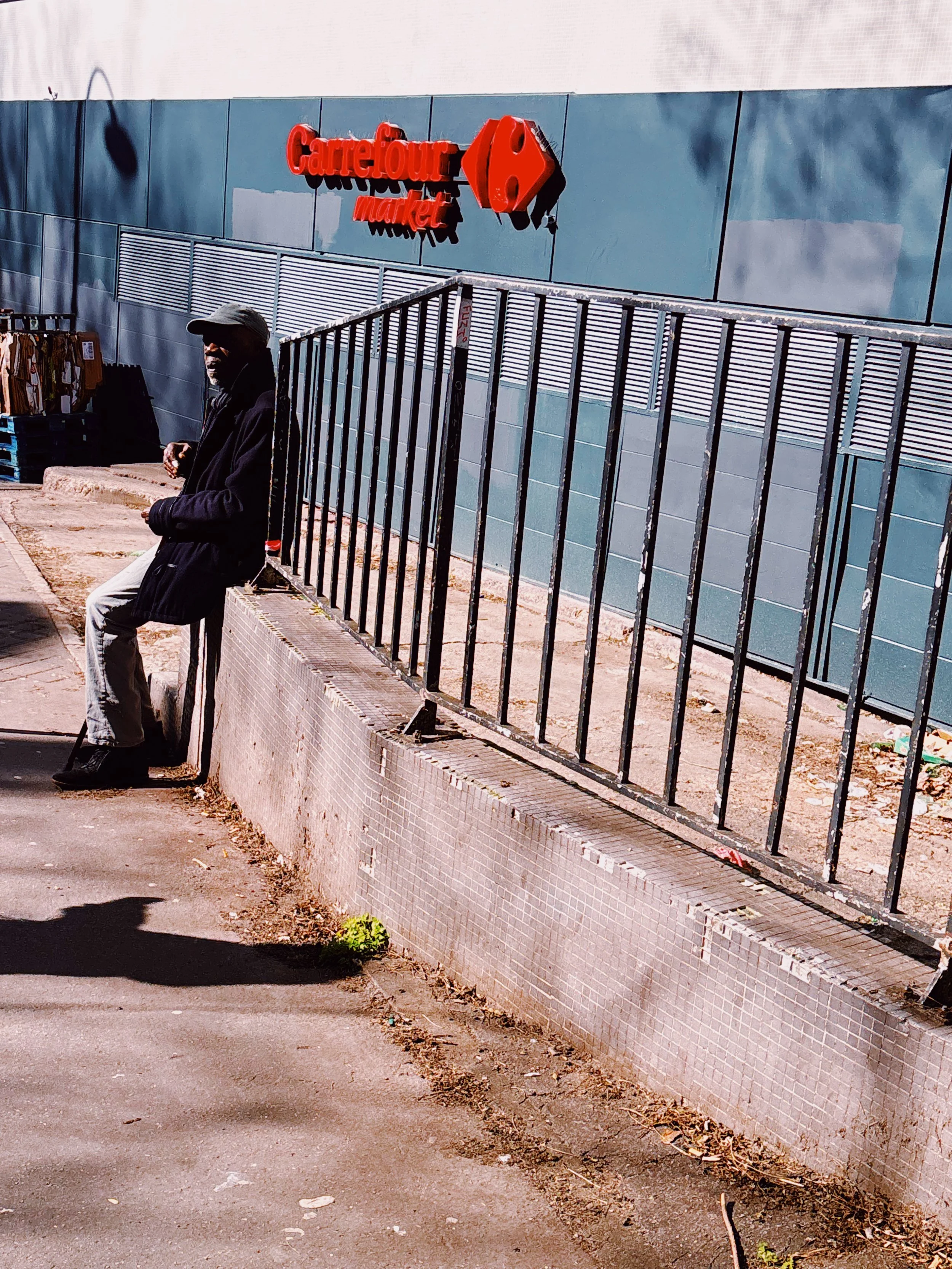 Vue d'une zone urbaine avec un filtre de station-service Carrefour Market, un homme en vêtements sombres, une barrière en métal noir et un trottoir abîmé avec des déchets.