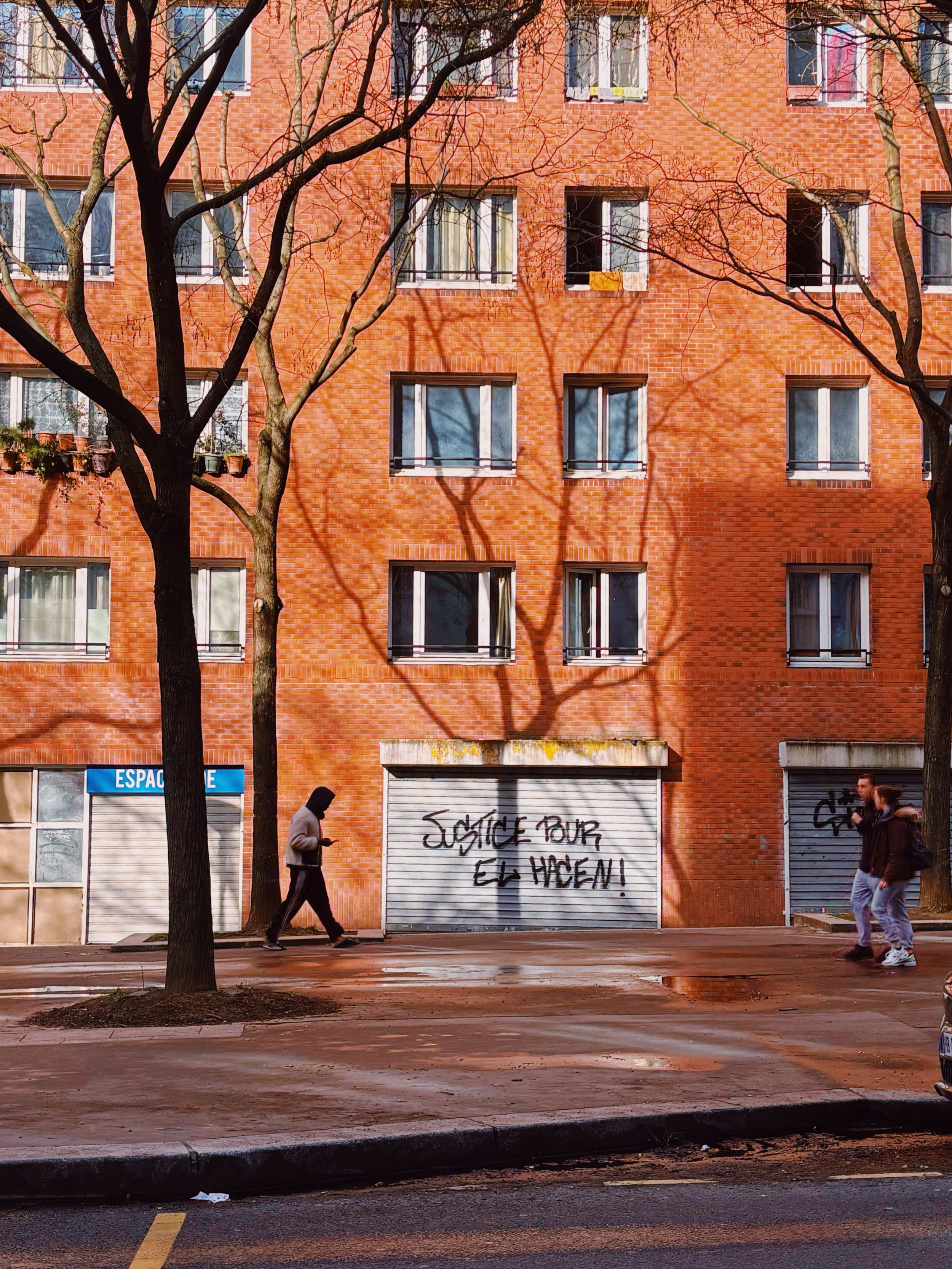 Façade d'un immeuble en briques rouges avec des fenêtres et des arbres sans feuilles projetant des ombres sur le mur. Deux personnes marchent devant, une avec un manteau beige et une autre avec un hoodie noir. Un panneau de graffiti dit : 'JUSTICE PO
