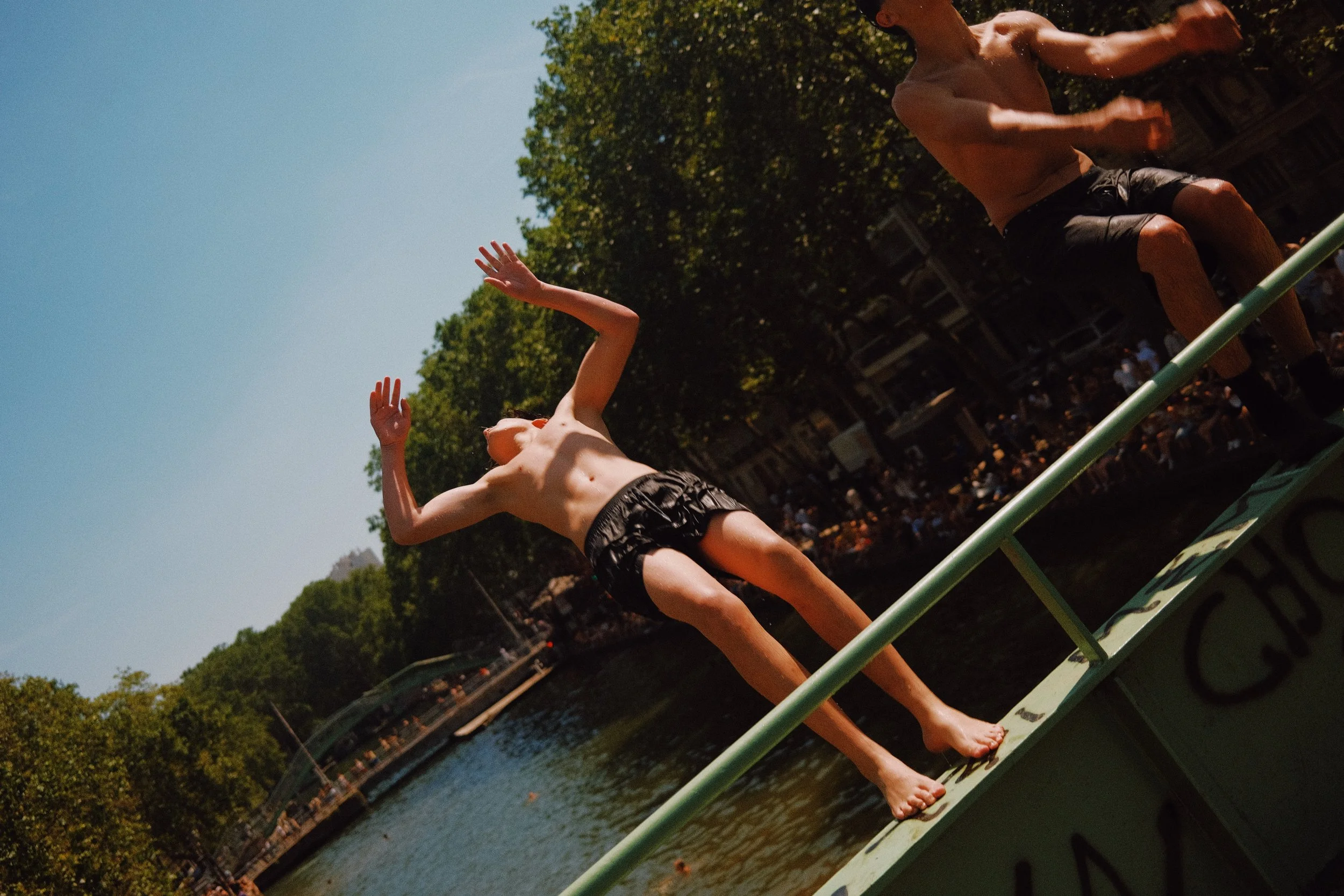 Deux personnes sautent dans l'eau en été, une d'elles a une peau claire et les autres ont une peau plus foncée, dans un parc avec des arbres verts et un corps d'eau, sous un ciel bleu. 
