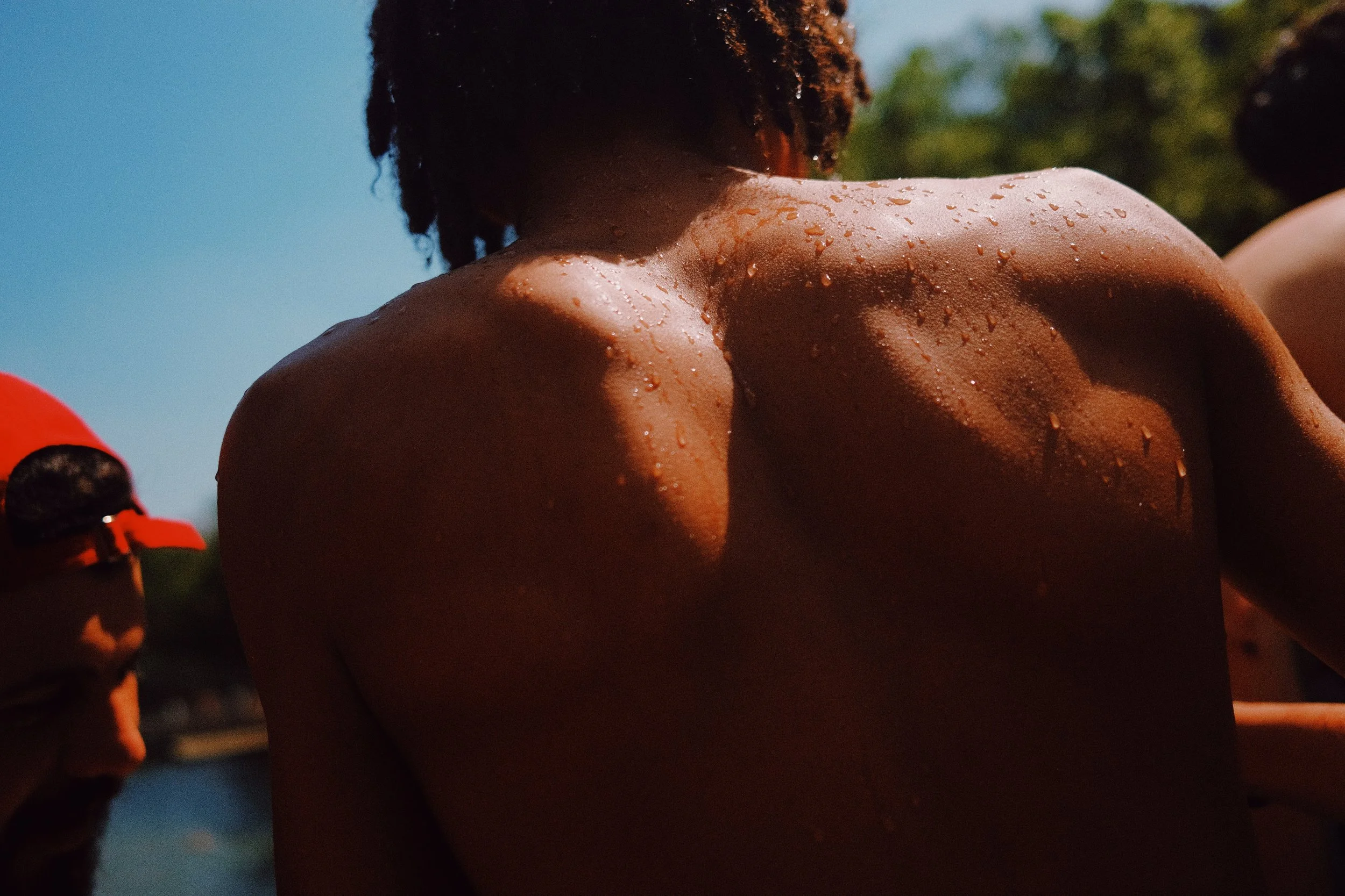 Une personne nue avec des gouttes d'eau sur la peau, vue de dos, avec un fond de ciel bleu et de verdure, lors d'une activité en plein air.