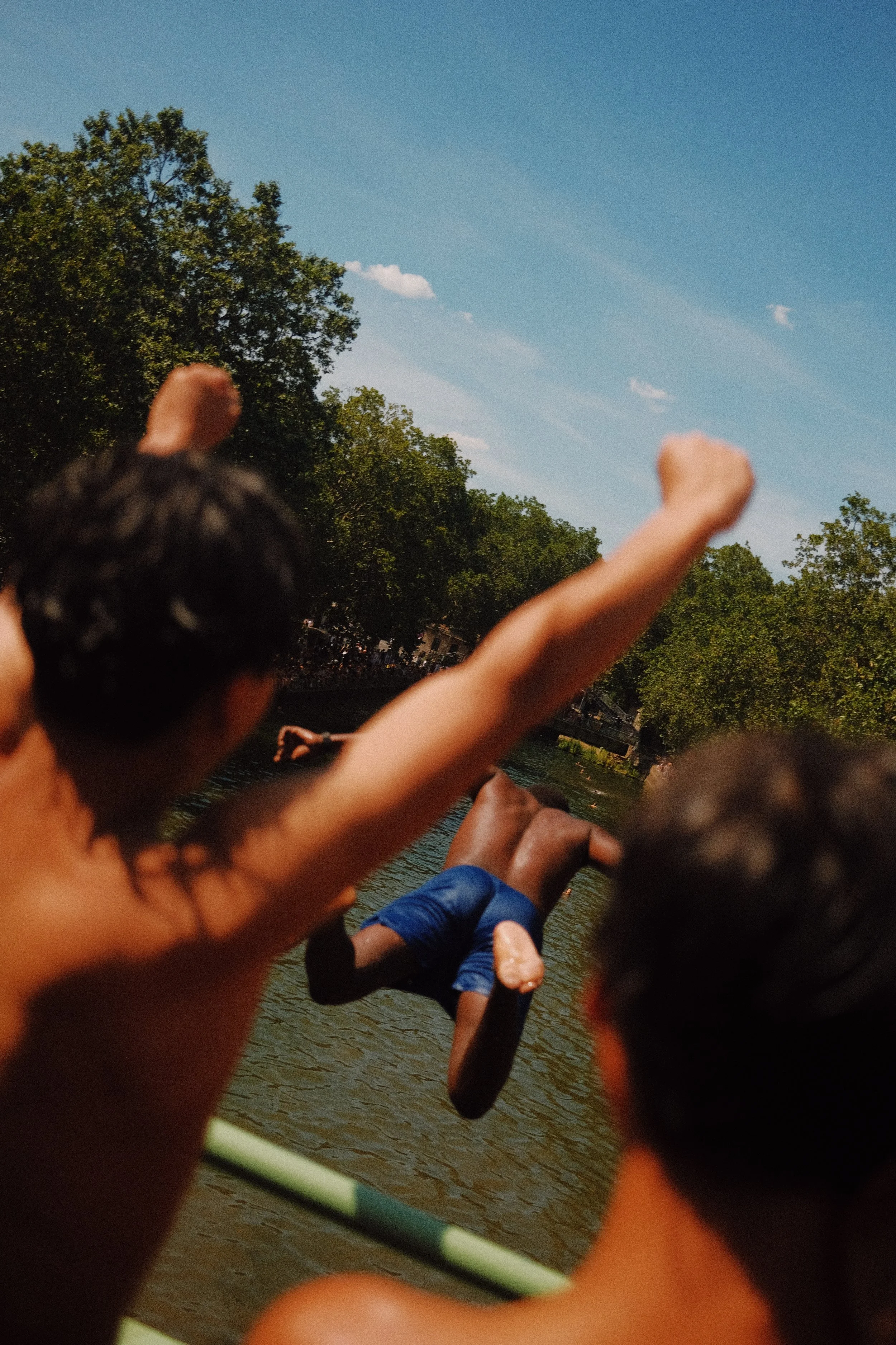 enfant nageant dans une rivière, entouré d'arbres, avec un groupe de personnes assises au bord de l'eau.