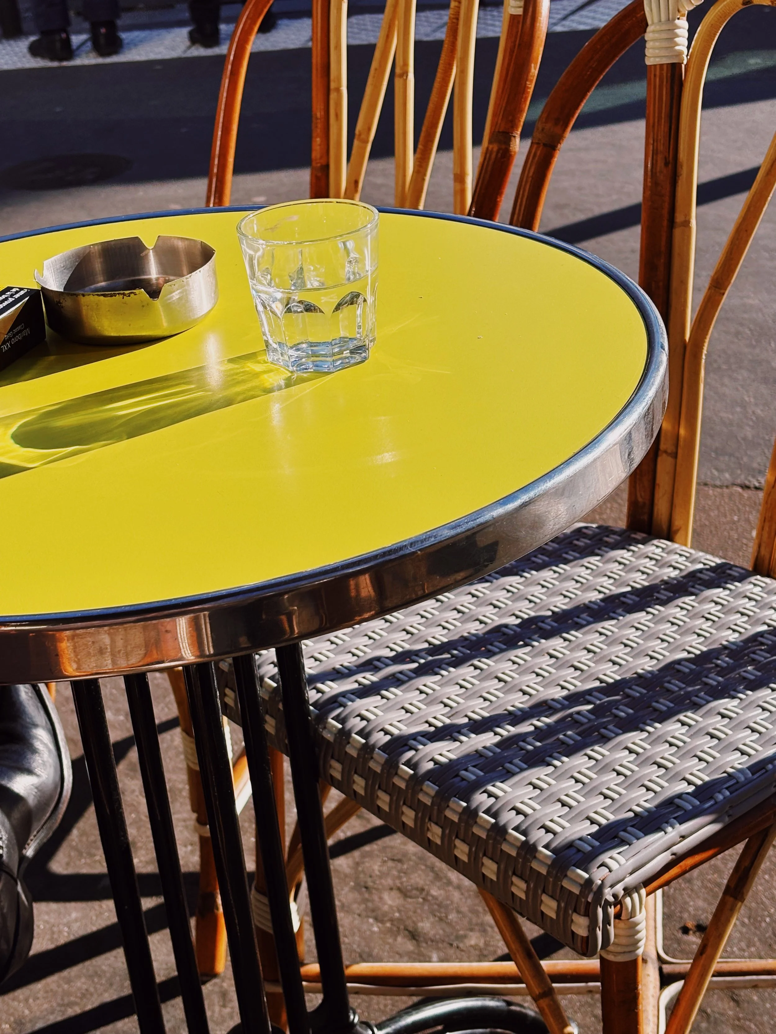 Verre d'eau et cendrier en métal sur une table jaune à l'extérieur, avec des chaises en bois et en rotin autour.