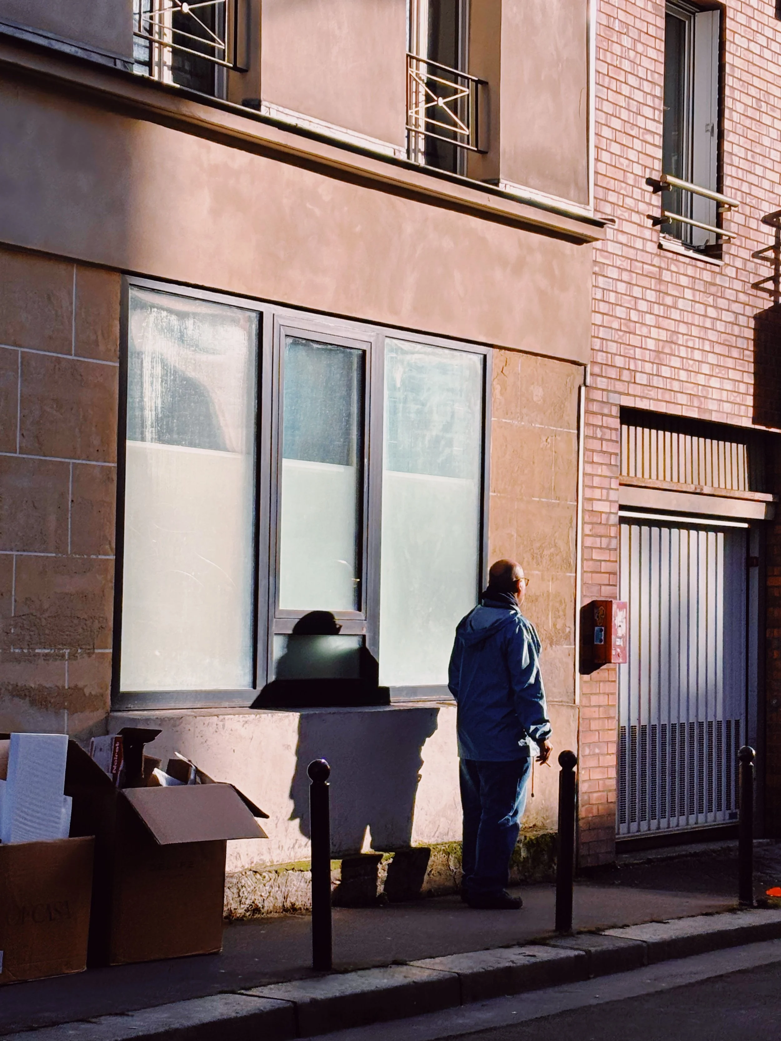 Deux hommes debout sur un trottoir, près d'une façade de bâtiment en pierre et briques, sous un lampadaire, avec des cartons en carton devant eux et un panneau d'incendie sur le mur.
