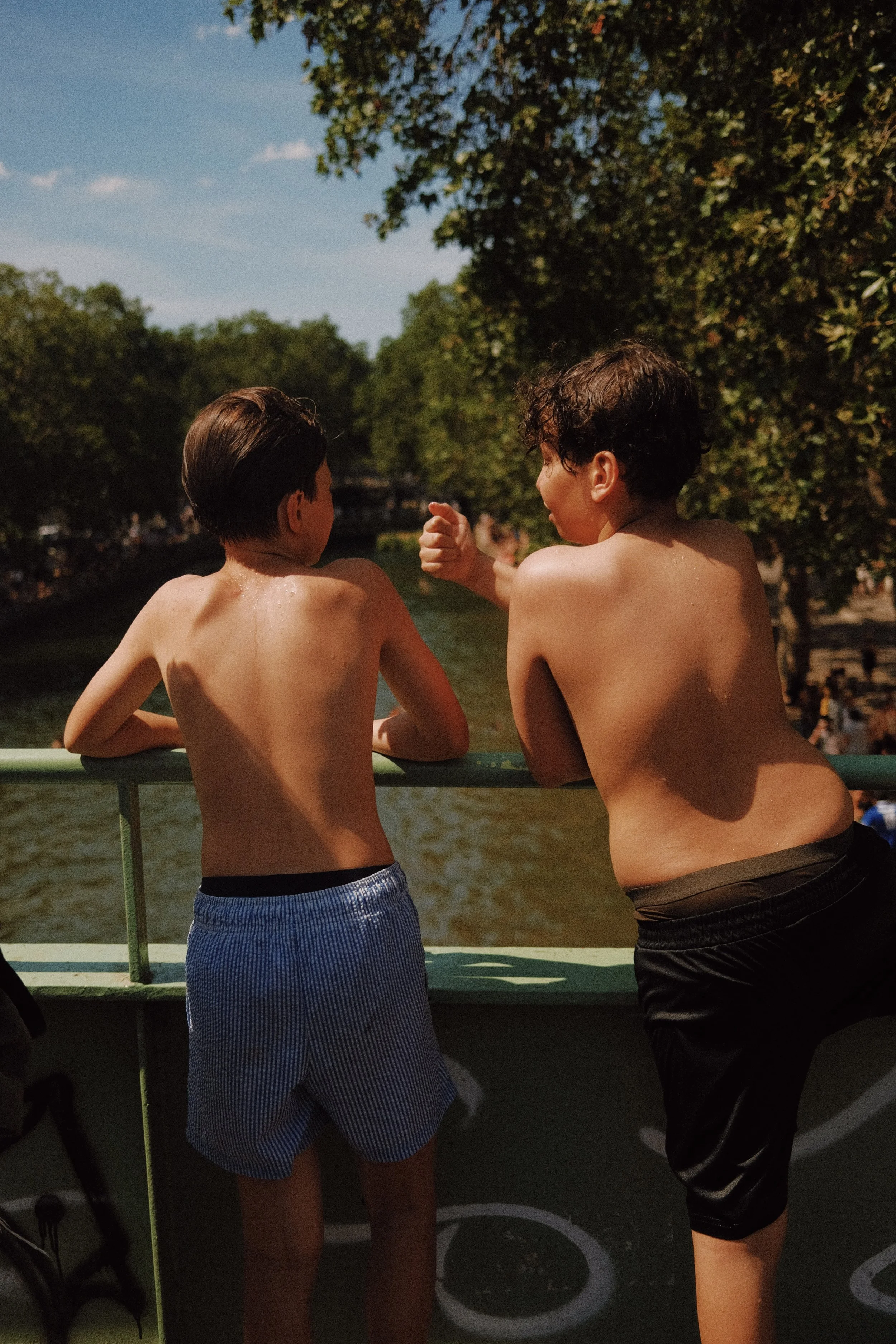 Deux garçon nus de dos, discutant ou jouant sur un bateau ou une plateforme à proximité d'un plan d'eau, avec des arbres en arrière-plan et un ciel bleu