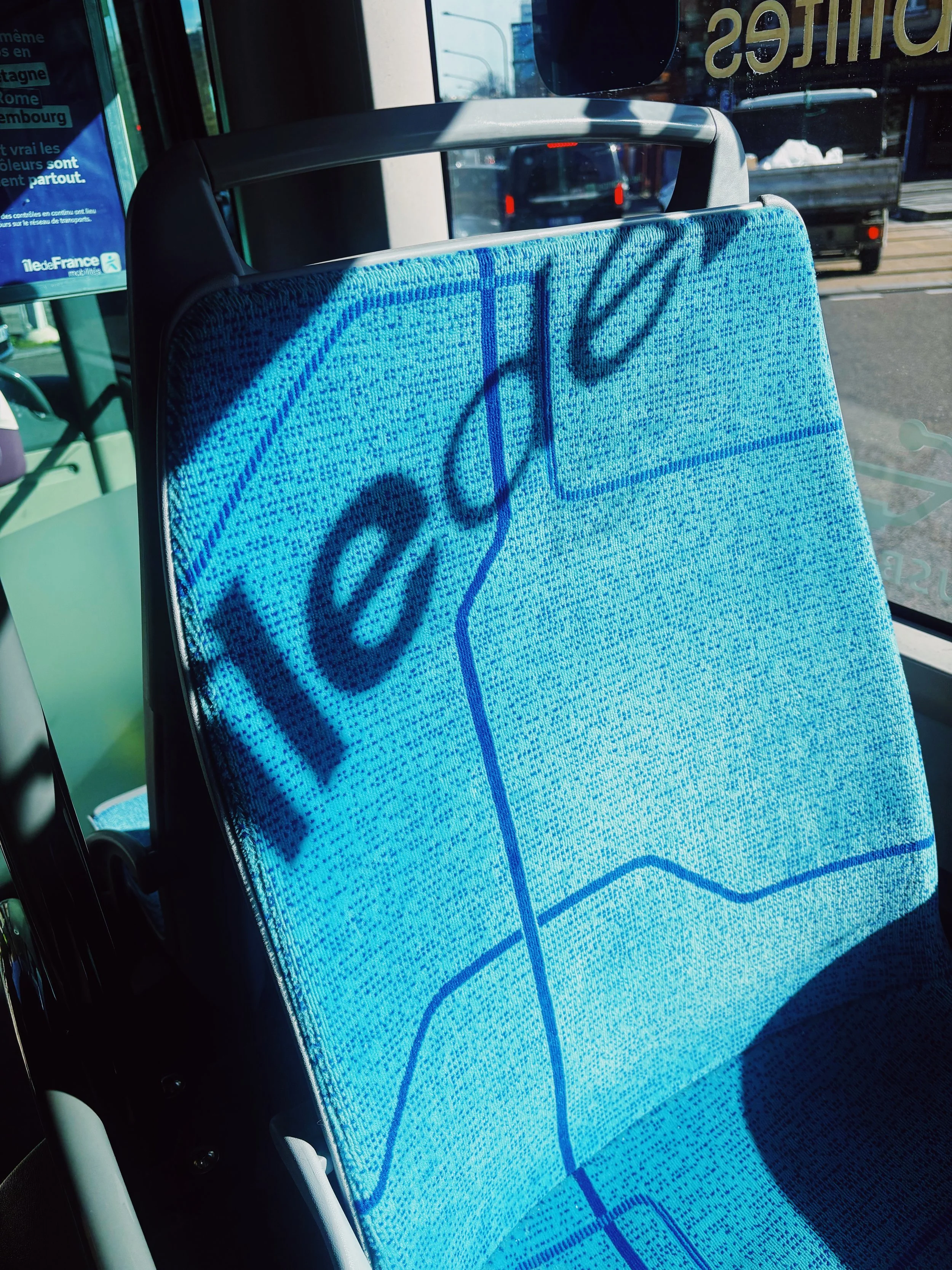 Assise dans un bus, une banquette rembourrée de bleu avec le nom 'Cali' écrit dessus, l'ombre du manche se projette sur la banquette, derrière la vitre, la route et un camion sont visibles.