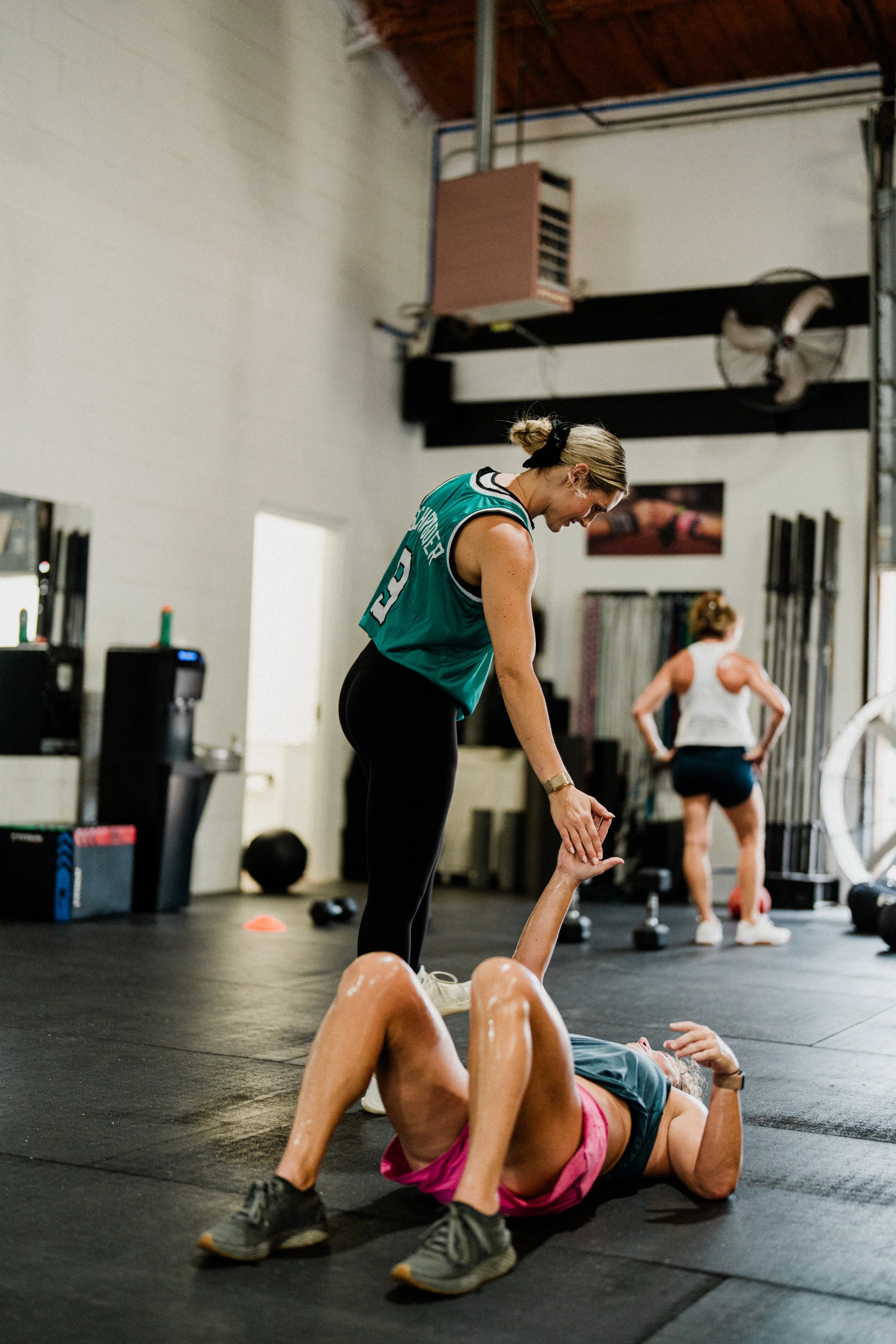 Women exercising in a gym, with one woman lying on the floor doing a leg raise and another woman standing, assisting her, while a third woman is seen in the background.