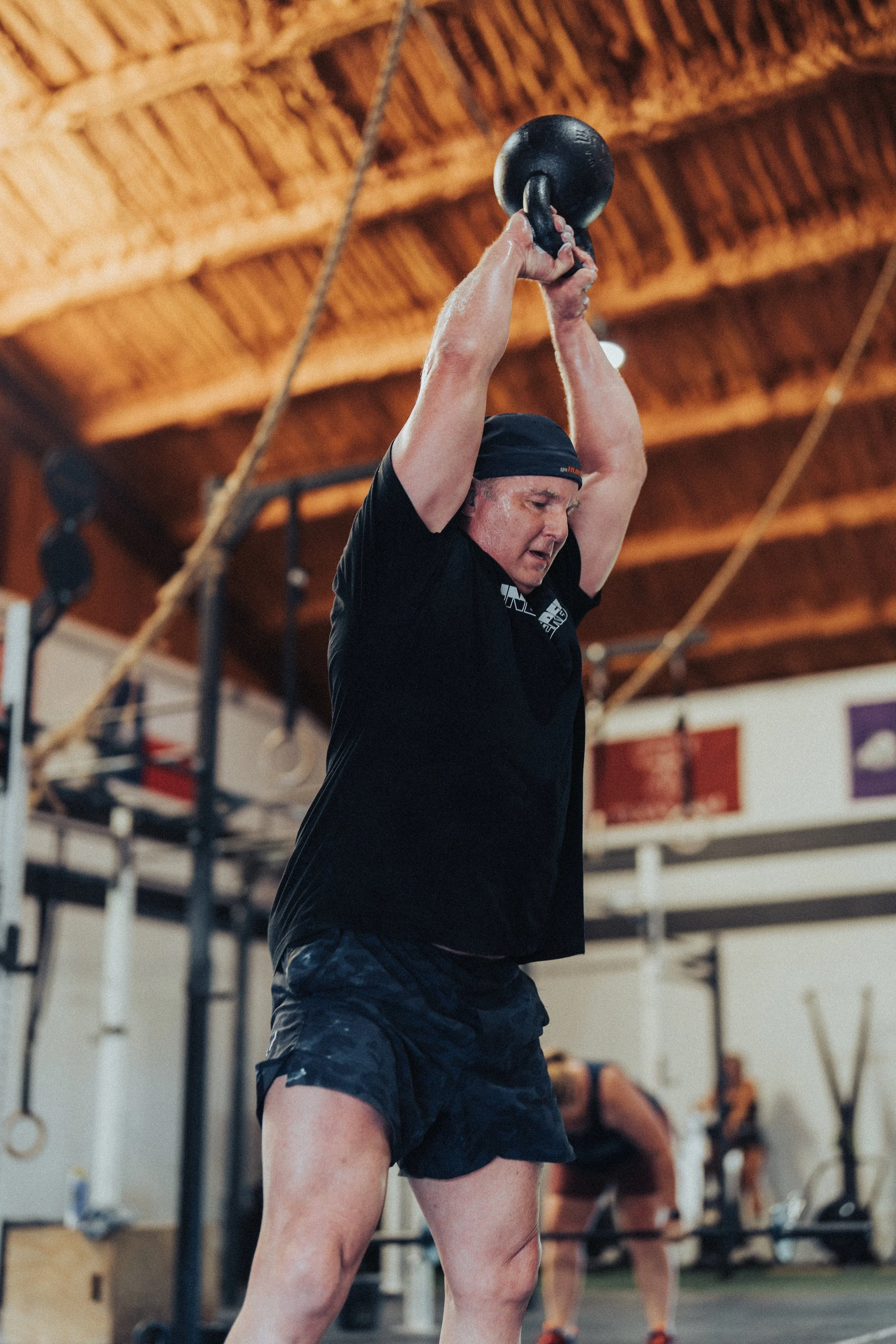 A man in athletic clothing and a headband lifting a black kettlebell overhead during a workout in a gym.