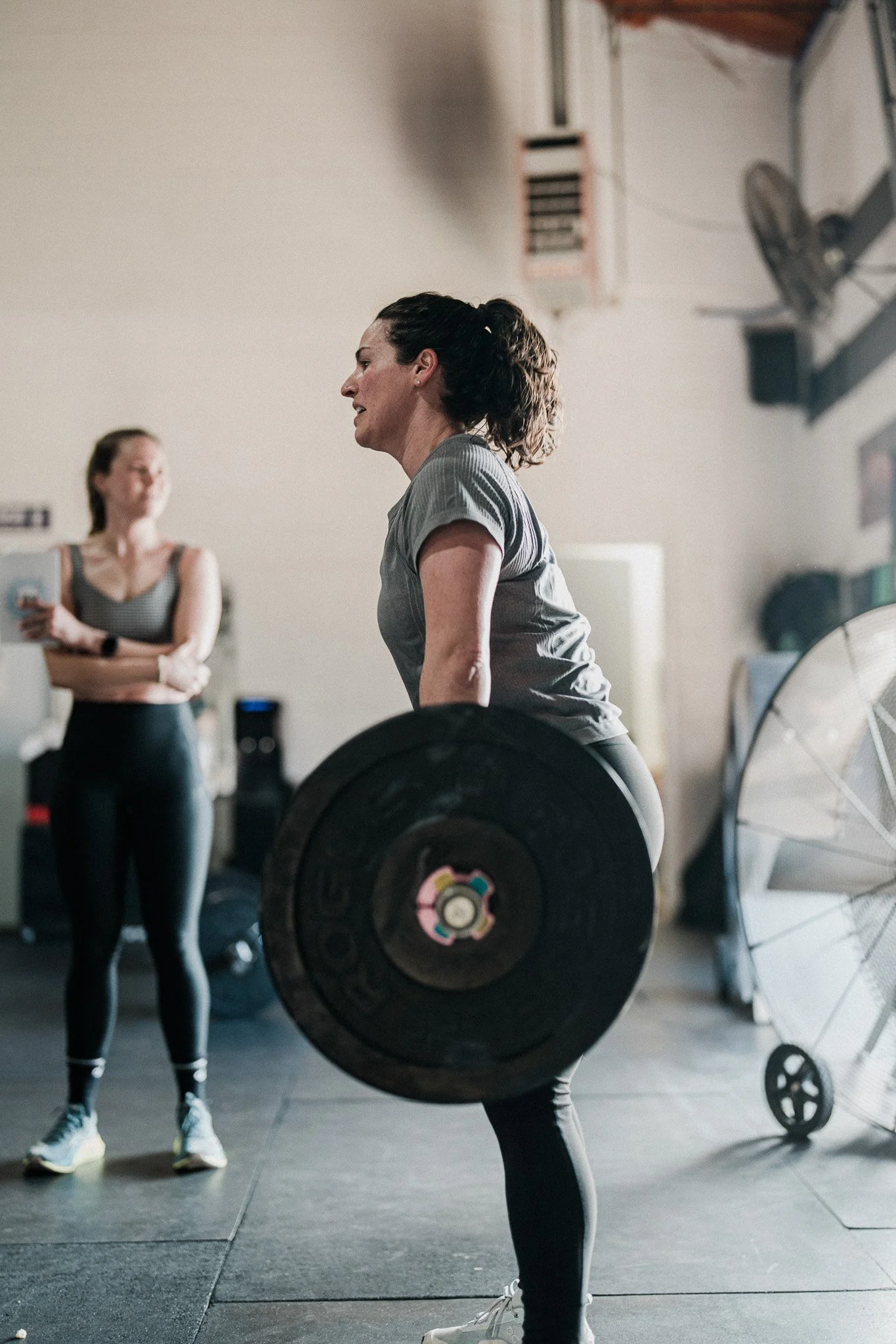 Two women in a gym lifting weights. The woman in the foreground is lifting a barbell and wearing a gray t-shirt and black leggings. The woman in the background is standing with her arms crossed, wearing a black and white sports bra and black pants.