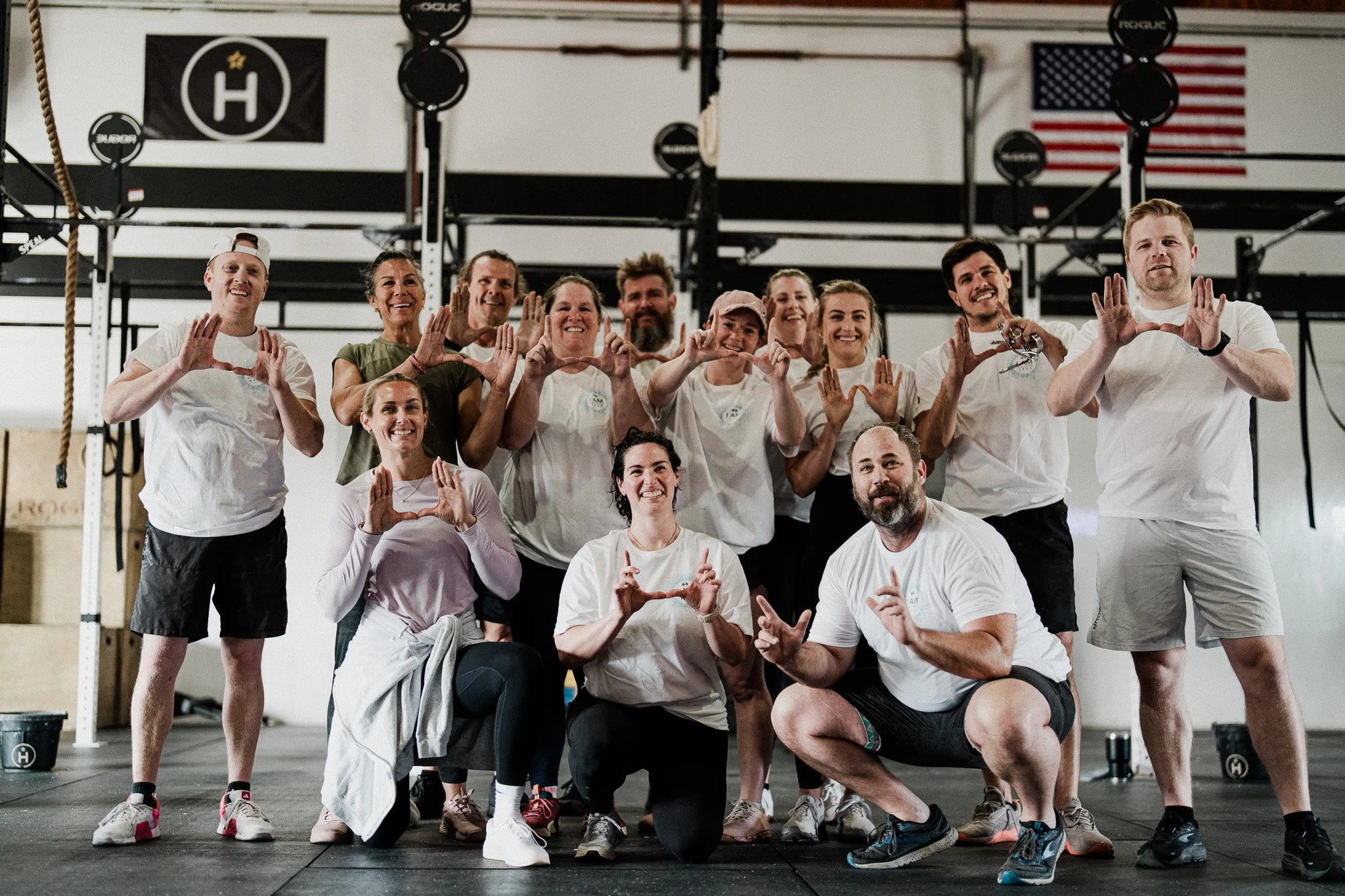 Group of people in a gym showing hand gestures to form hearts and peace signs, with gym equipment and American and black flags in the background.