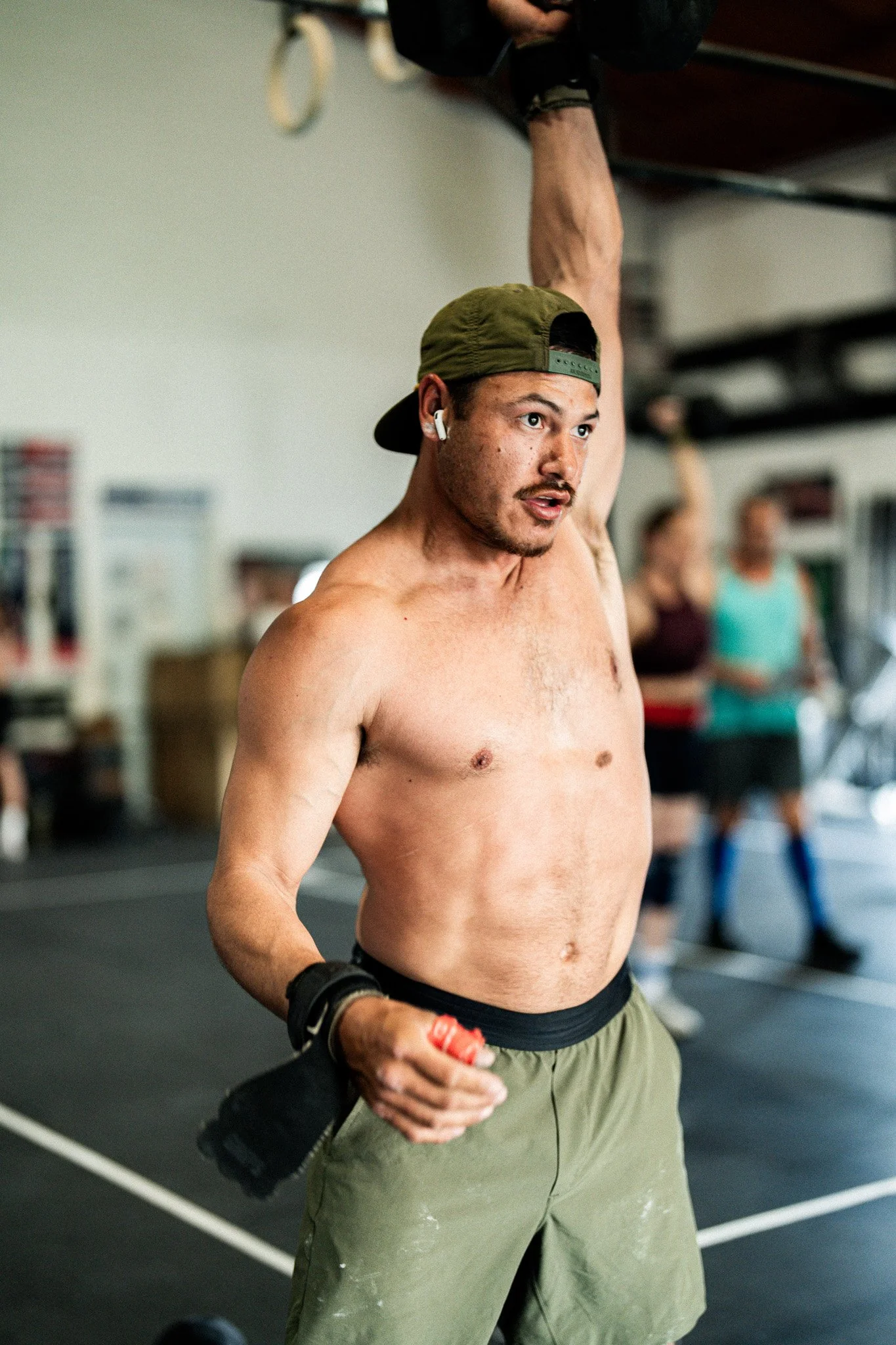 A shirtless man wearing a green baseball cap backward, green shorts, and wireless earbuds, lifting one arm in a fitness gym. In the background, other people are exercising.