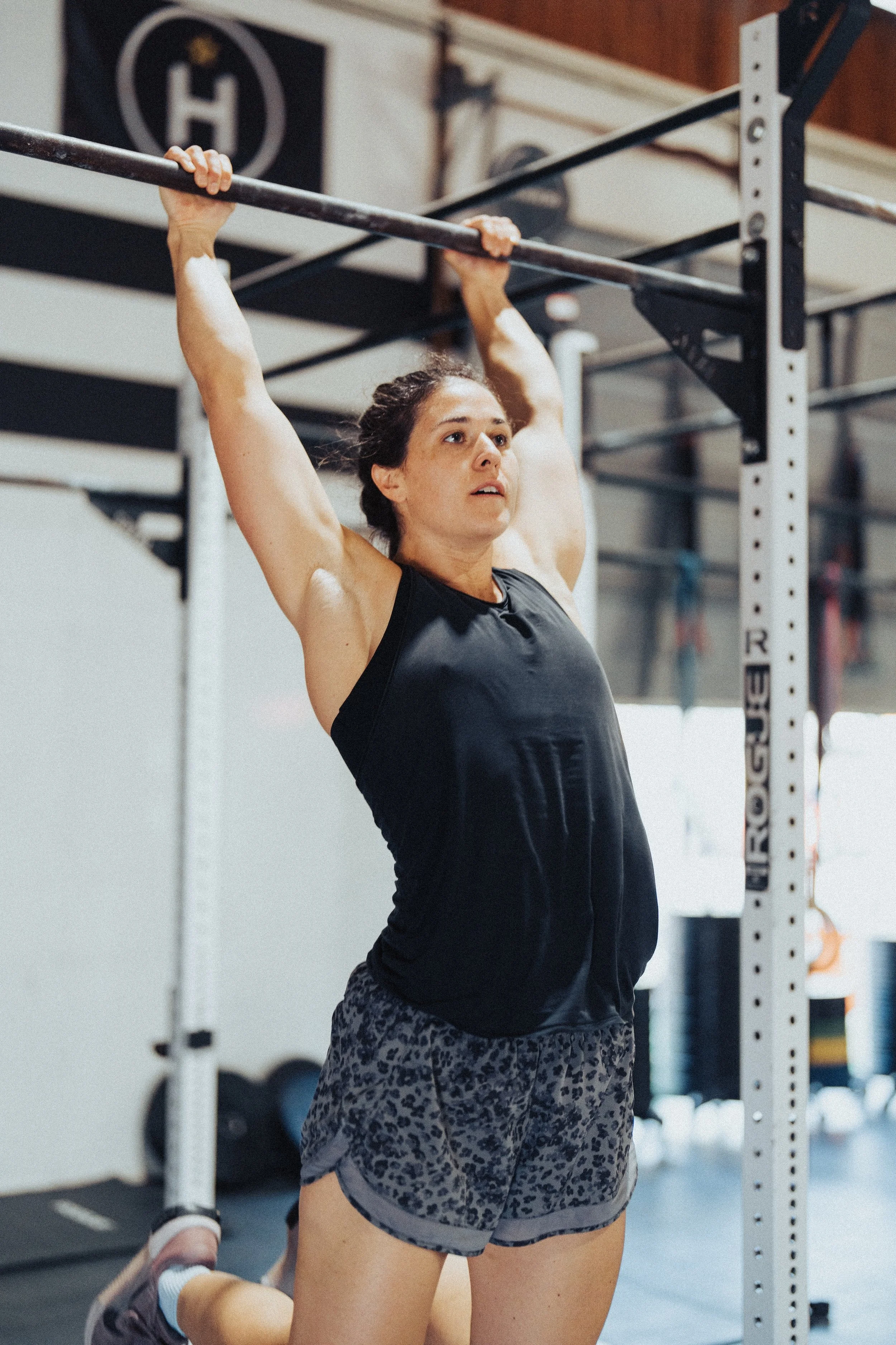 A woman wearing a black tank top and leopard print shorts is doing a pull-up exercise in a gym, using a pull-up bar attached to a metal frame.