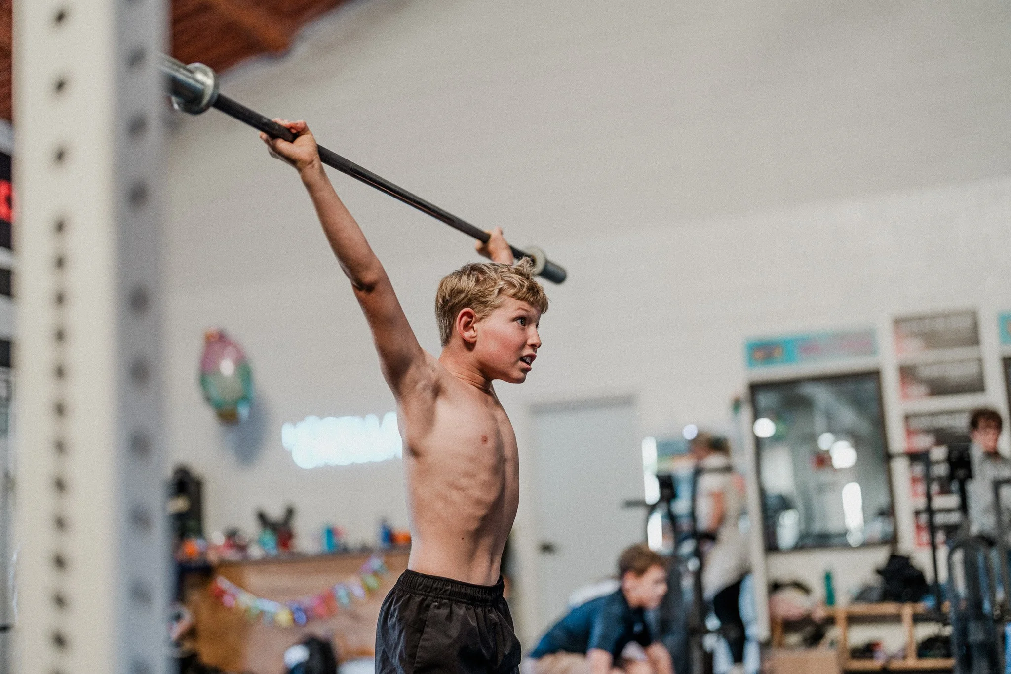 A young boy with blonde hair and no shirt is lifting a barbell overhead at a gym, with a focused expression.