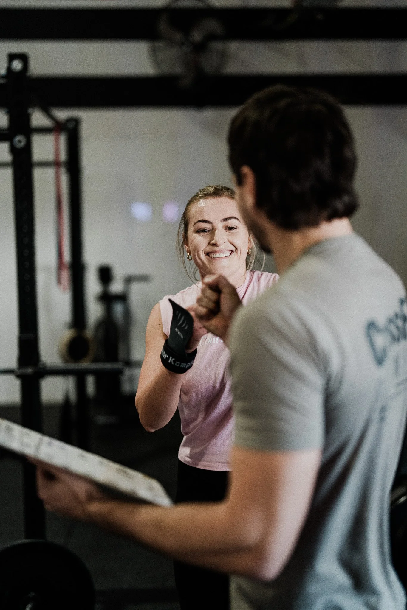A woman smiling and talking to a man during a workout session at a gym. The woman is wearing a pink tank top and gloves, and the man is wearing a gray t-shirt. They are standing near gym equipment.