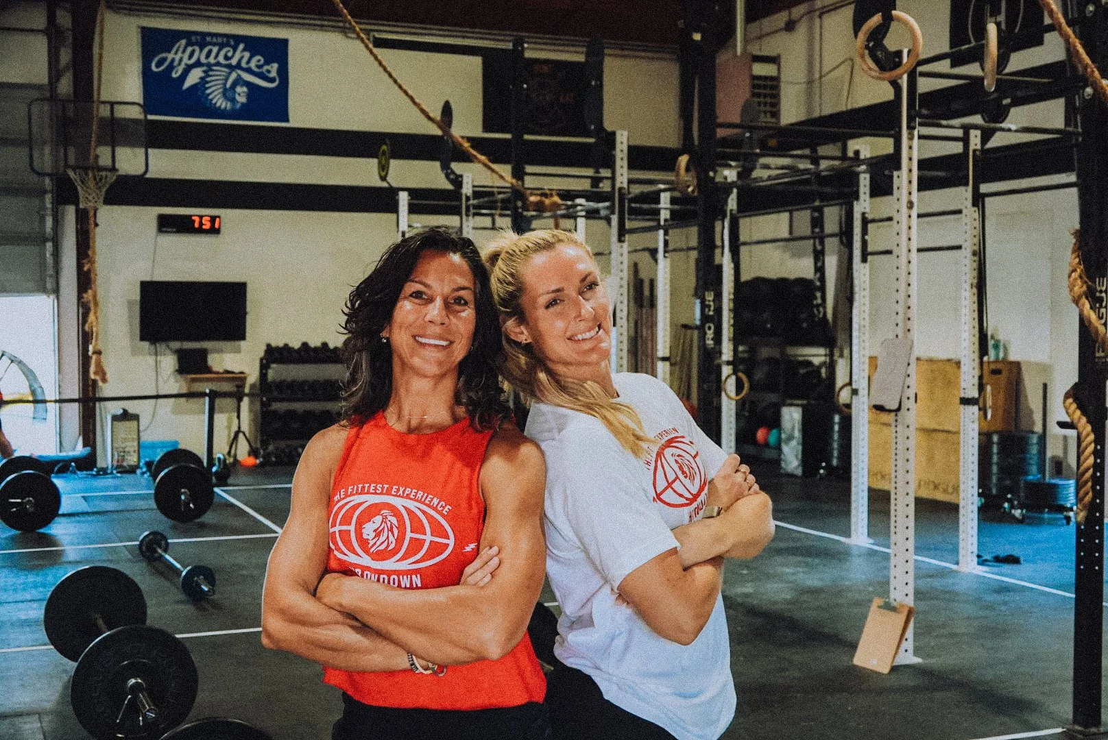 Two women standing in a gym, smiling with arms crossed, wearing athletic shirts with a lion logo, surrounded by gym equipment like barbells, weights, and gymnastic rings.