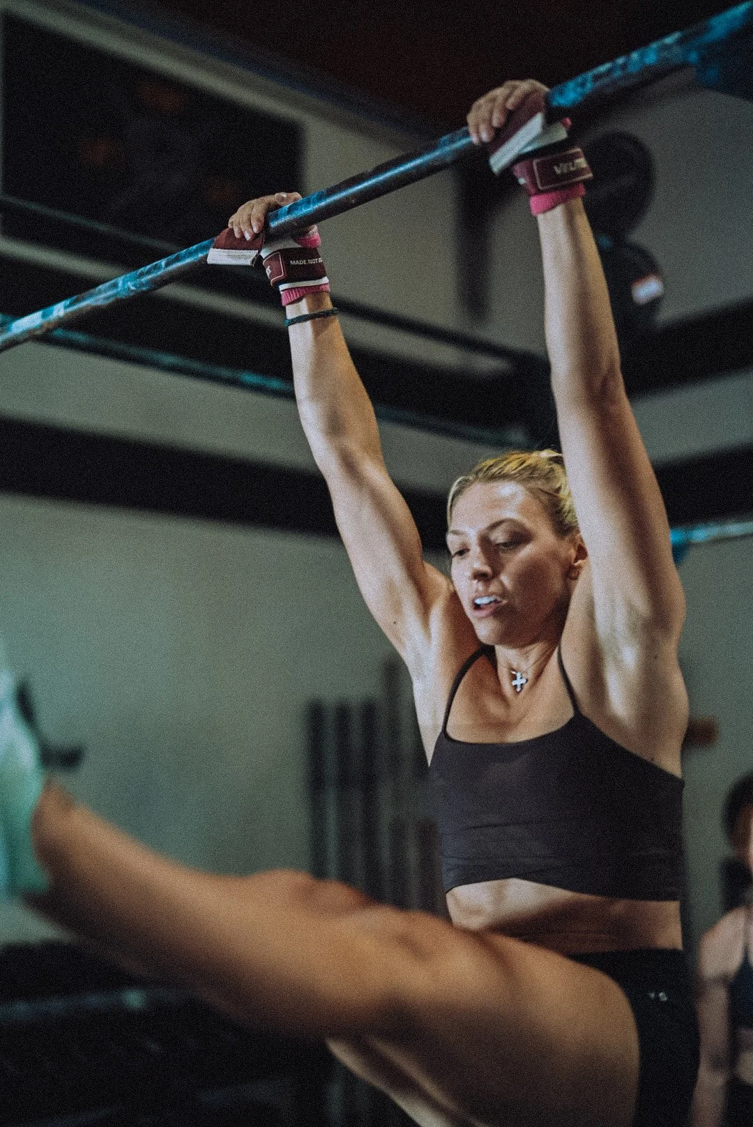 A woman with blonde hair in a ponytail wearing a black sports bra and wrist wraps, hanging from a pull-up bar in a gym.