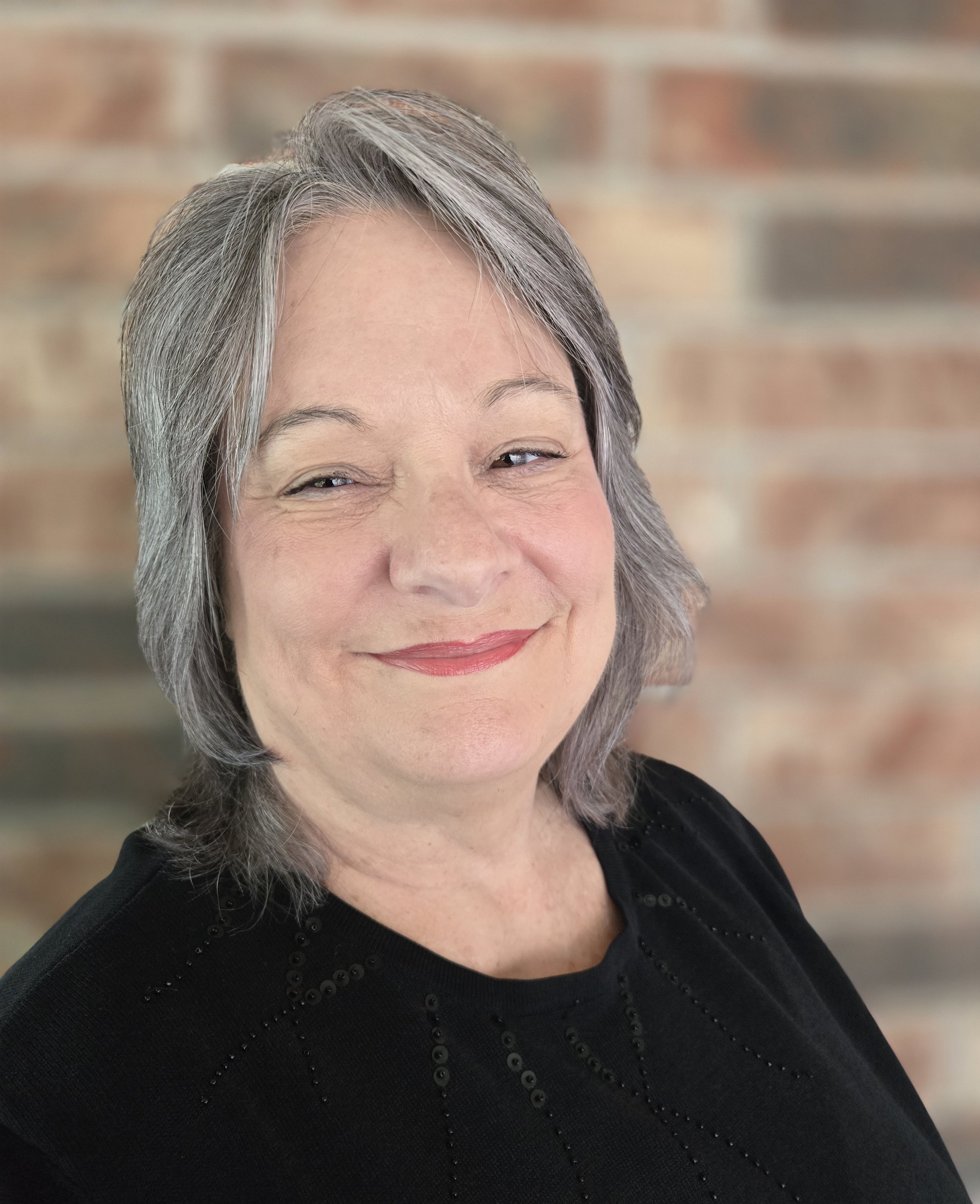 A smiling woman with short gray hair wearing a black top standing in front of a blurred brick wall.