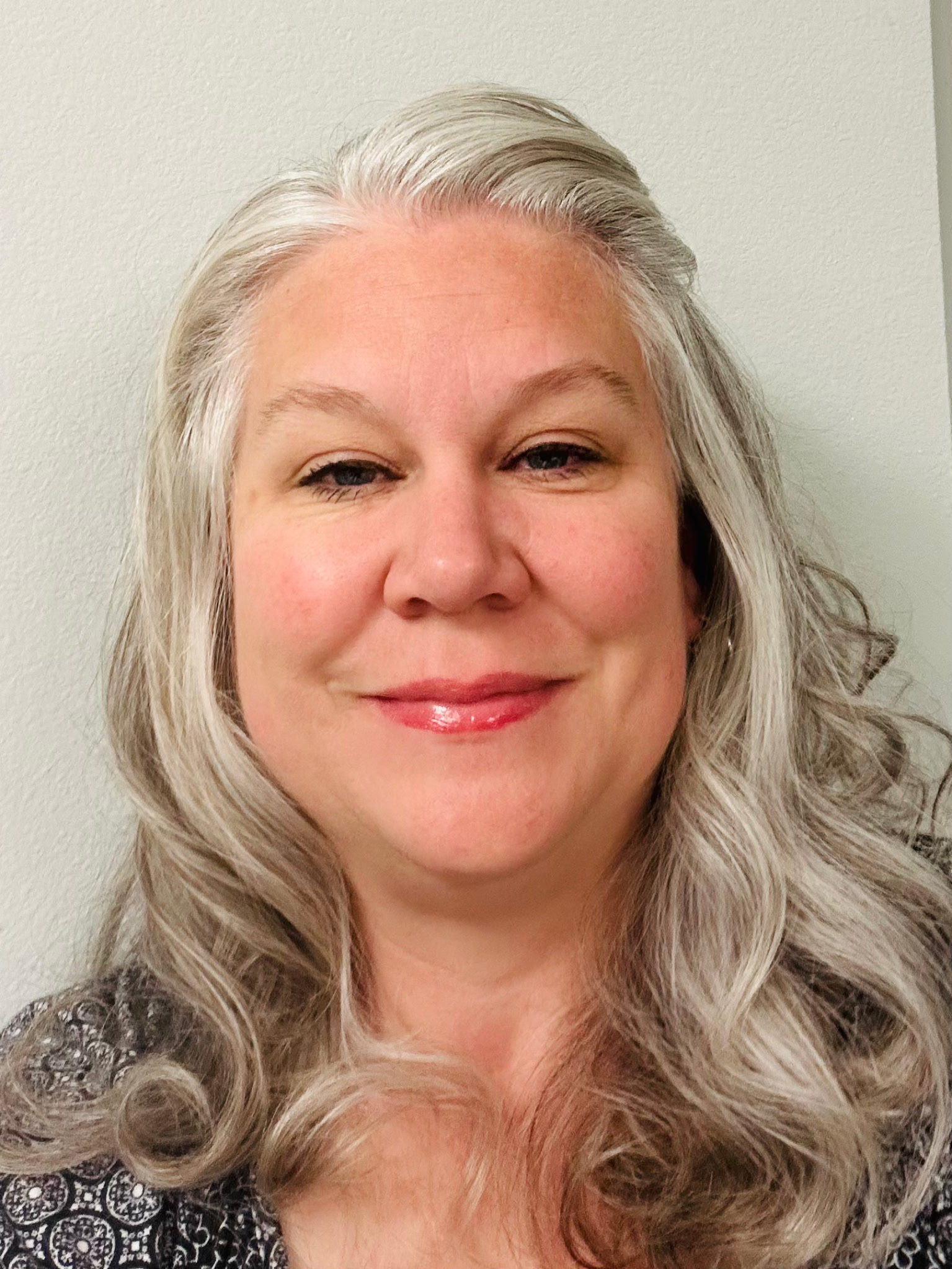 A close-up photo of a woman with blonde, wavy hair and light makeup, standing in front of a plain light-colored wall.
