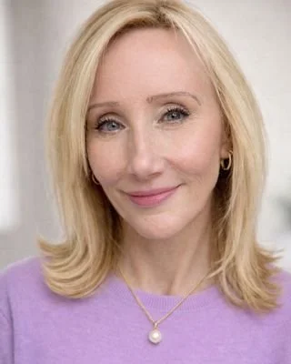 A woman with blonde hair smiling, wearing a purple top and pearl necklace.