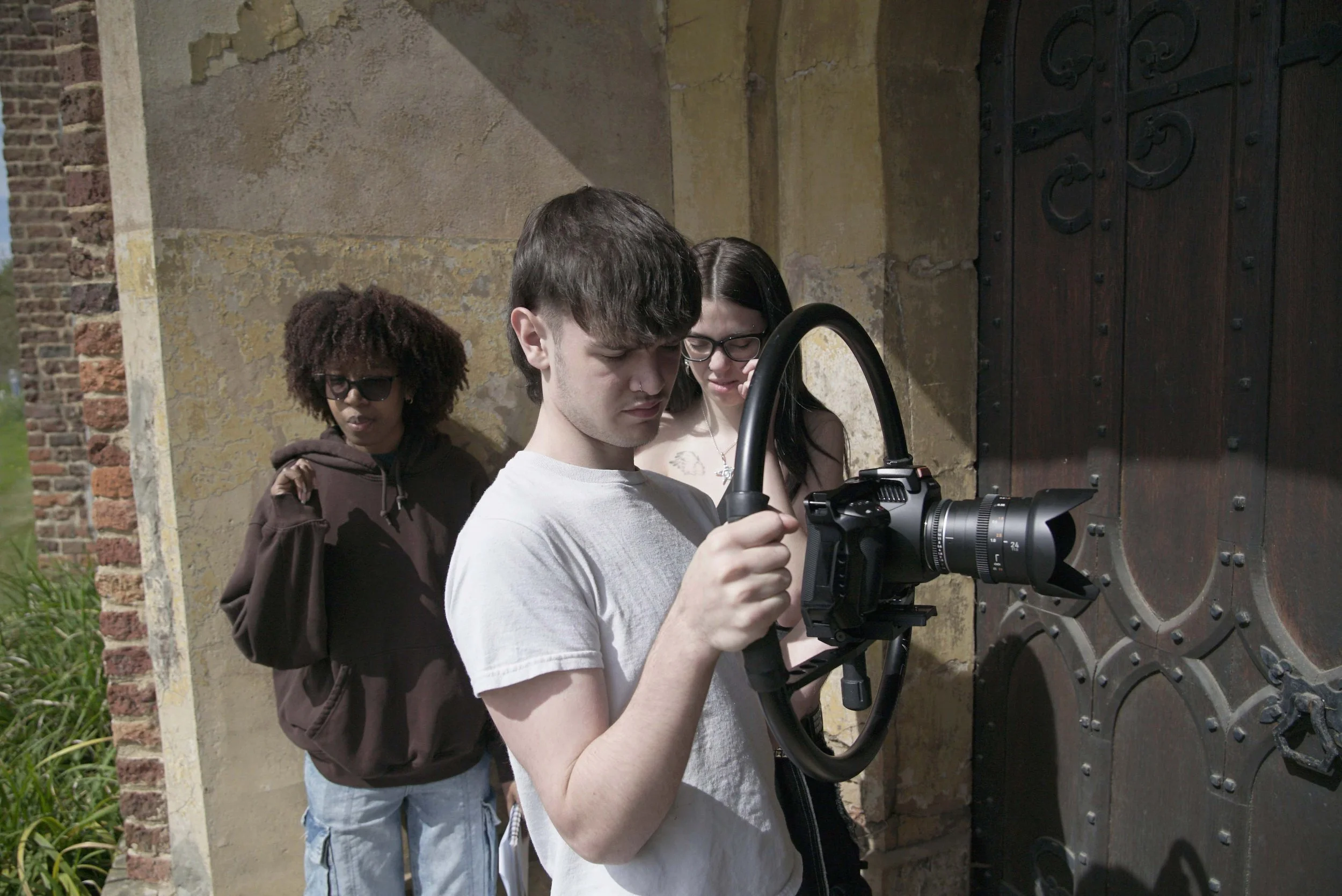 Three young people are gathered outdoors, with two women and one man, examining a professional camera mounted on a tripod in front of an old, textured metal door, on a sunny day.