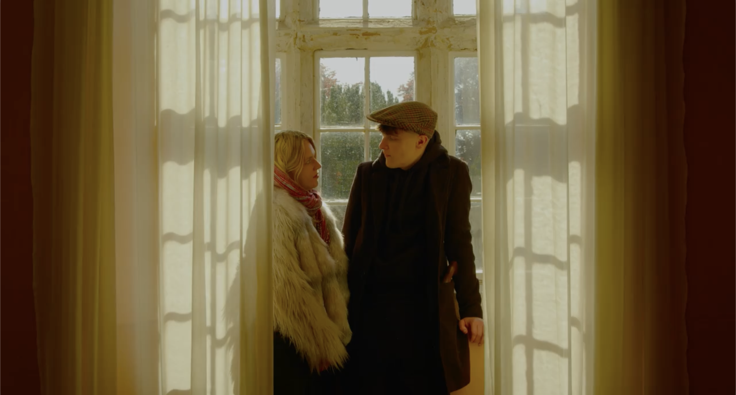 A man and woman standing close together near a window with cream-colored curtains, indoors during daytime.