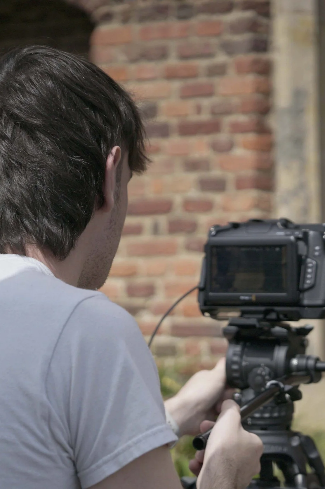 A person with brown hair wearing a white t-shirt is shooting with a camera mounted on a tripod, outdoors in front of a brick wall.