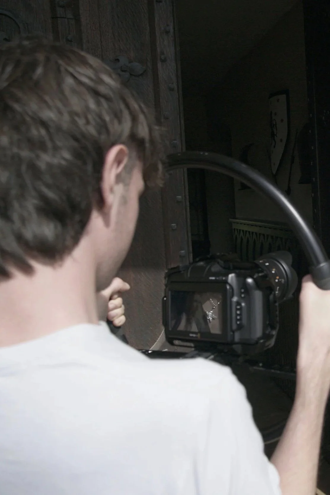 A person with brown hair operating a camera with a large lens and a screen, aiming it towards a dark area, possibly a door or passage.