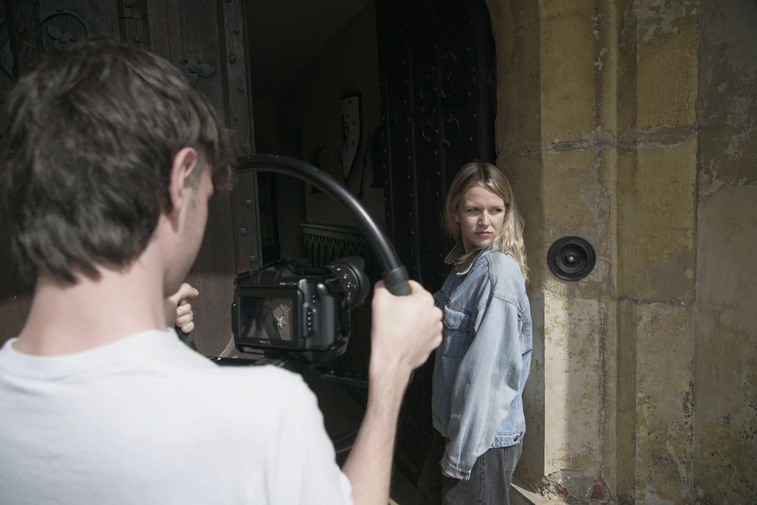 A woman with blonde hair and a denim jacket poses outdoors, while a man with a camera photographs her against a weathered brick wall.