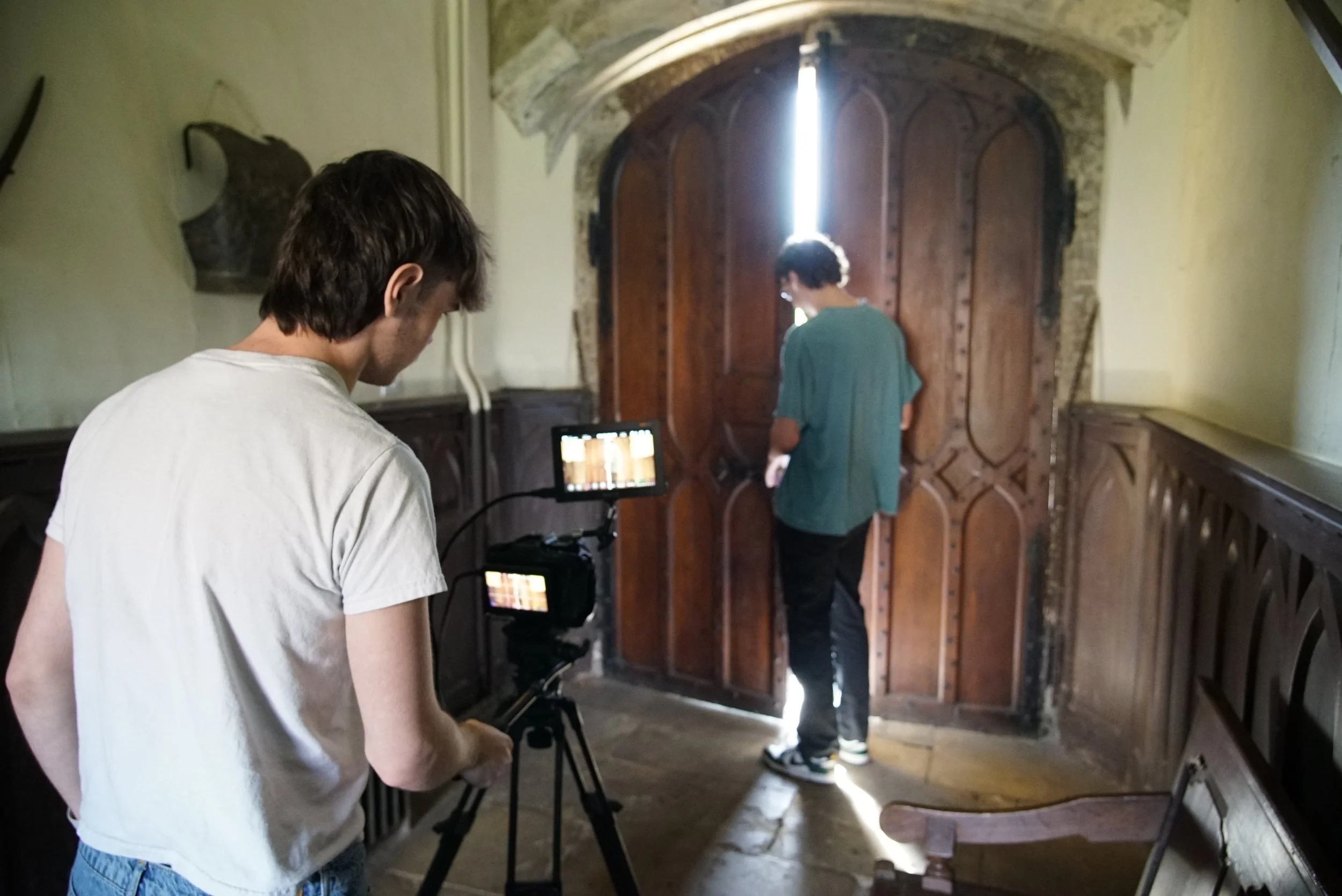 Two people filming a person standing in front of a large wooden door with stone archway. One person is looking at a monitor, and the other is standing near the door as if performing or acting.