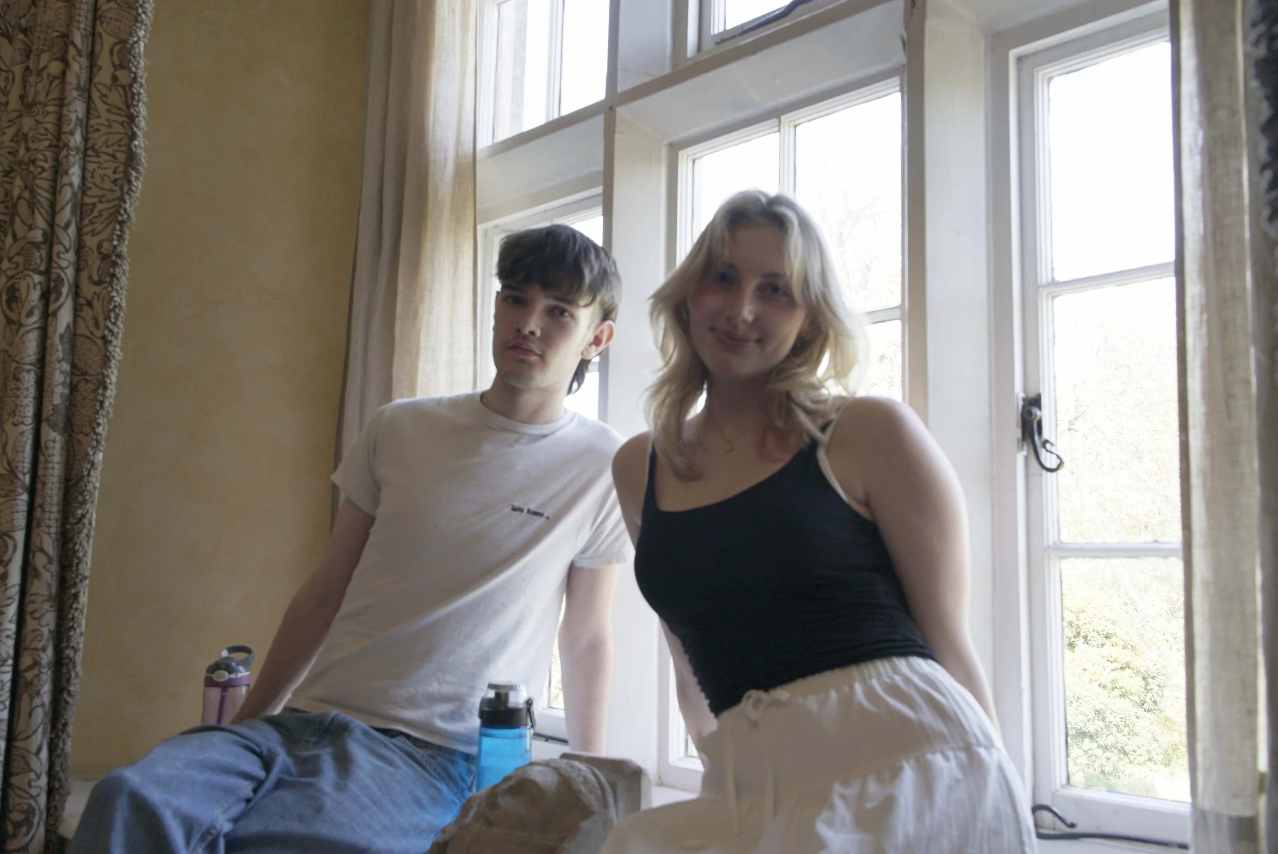 A young man and woman standing close together in front of a large window in a room with beige walls and patterned curtains, smiling at the camera.