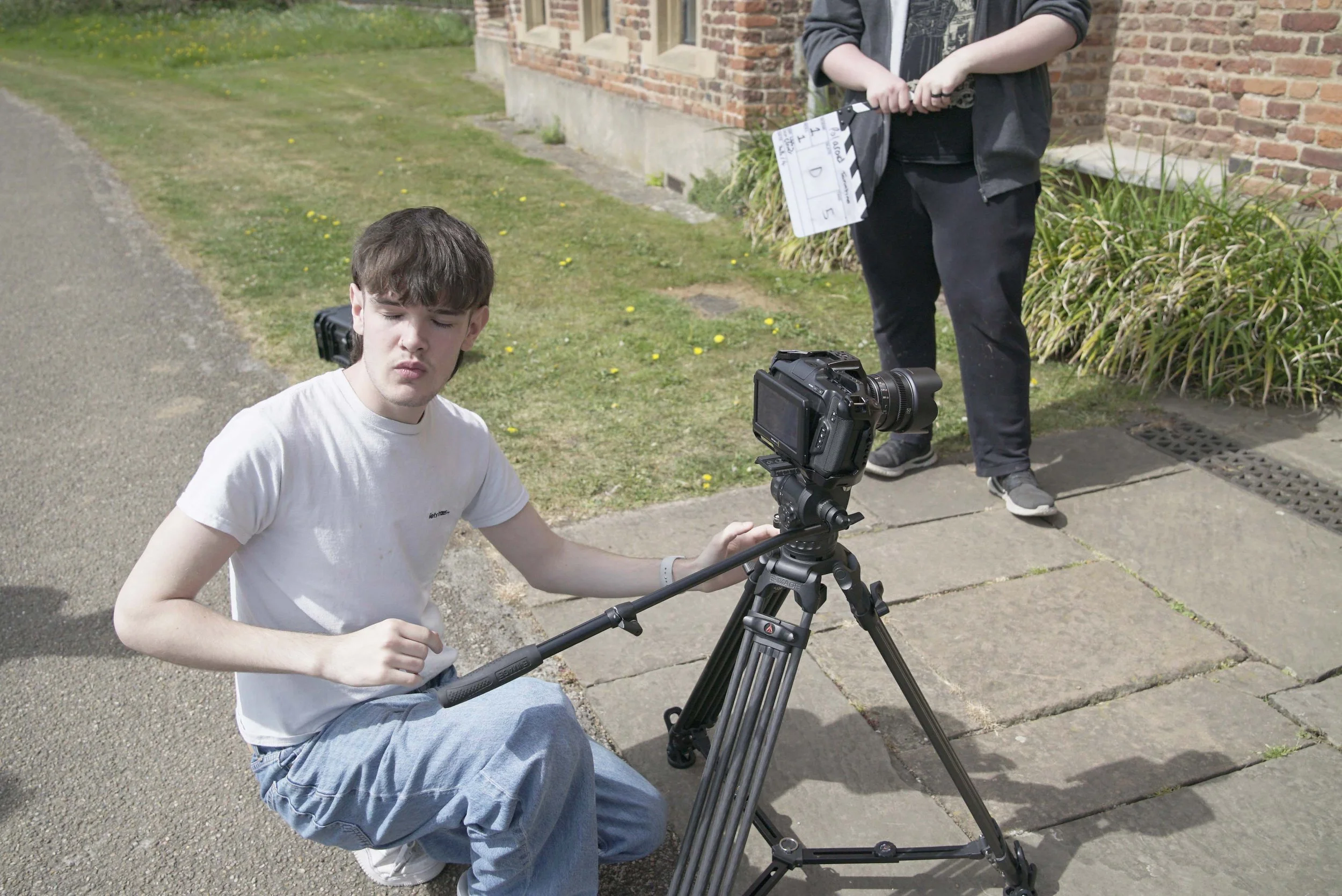 A young man with brown hair and a white t-shirt operating a professional video camera on a tripod outdoors, with a person holding a clapperboard standing near a brick building in the background.