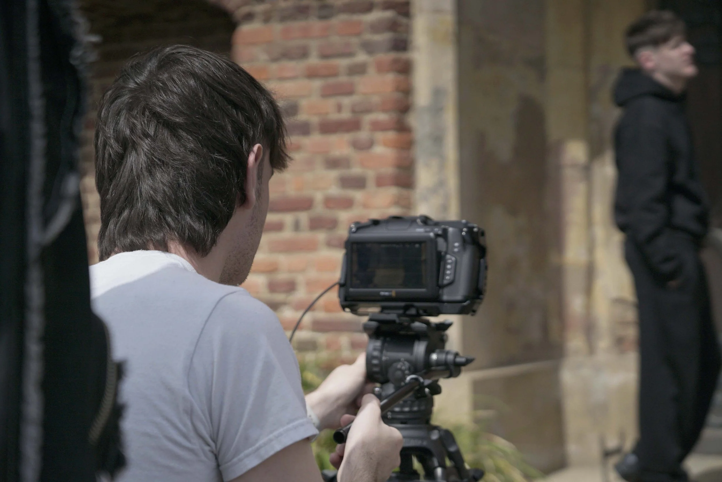 A person with brown hair is filming with a professional camera on a tripod in front of a brick wall, with another person standing nearby, wearing a black hoodie.