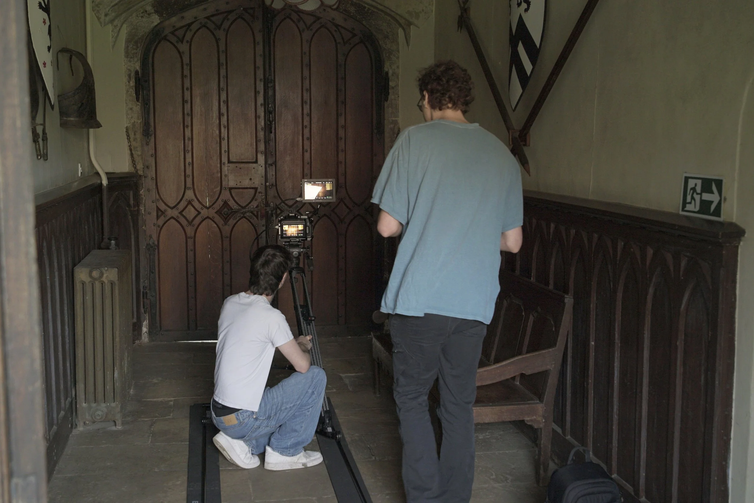Two individuals setting up camera equipment in a historic room with dark wooden doors and stone walls, one kneeling and one standing, amid various accessories and a directional emergency exit sign on the wall.