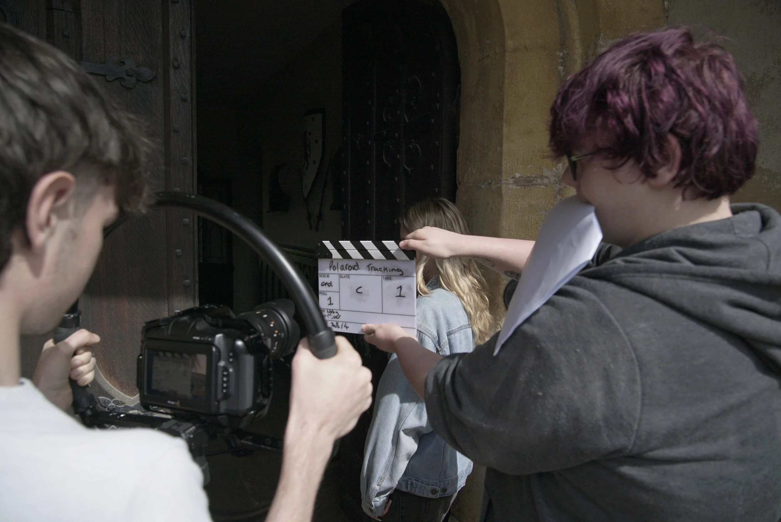 Crew filming a scene for Polaroid tracking in a rustic indoor setting with stone walls. Crew members hold a clapperboard and operate a camera.