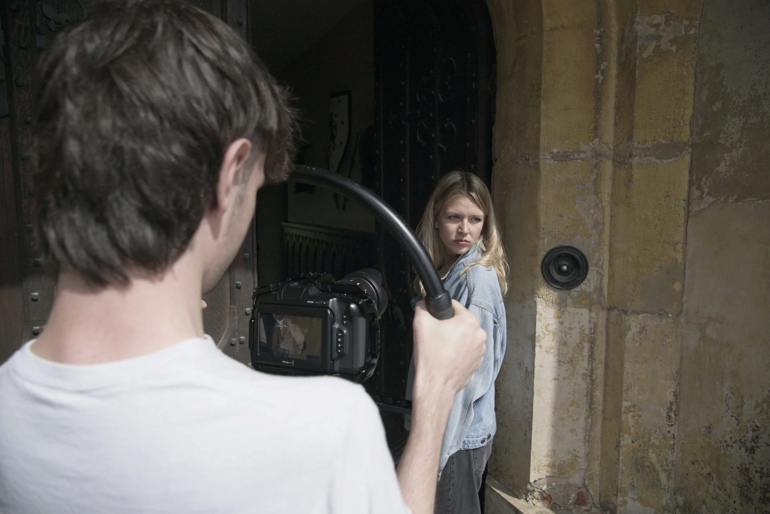 Photographer filming a young woman with blonde hair in a denim jacket against an aged yellow brick wall.