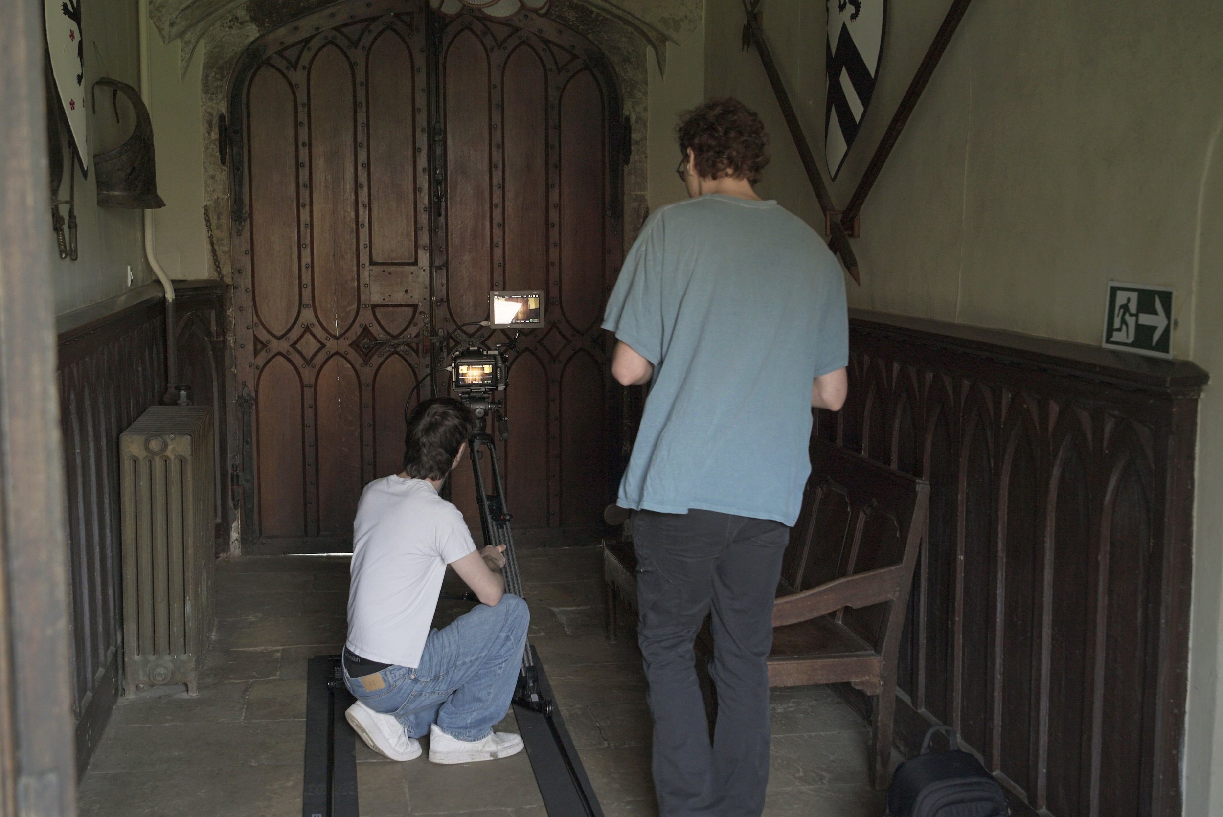 Two people setting up camera equipment in front of a large wooden door inside a building.