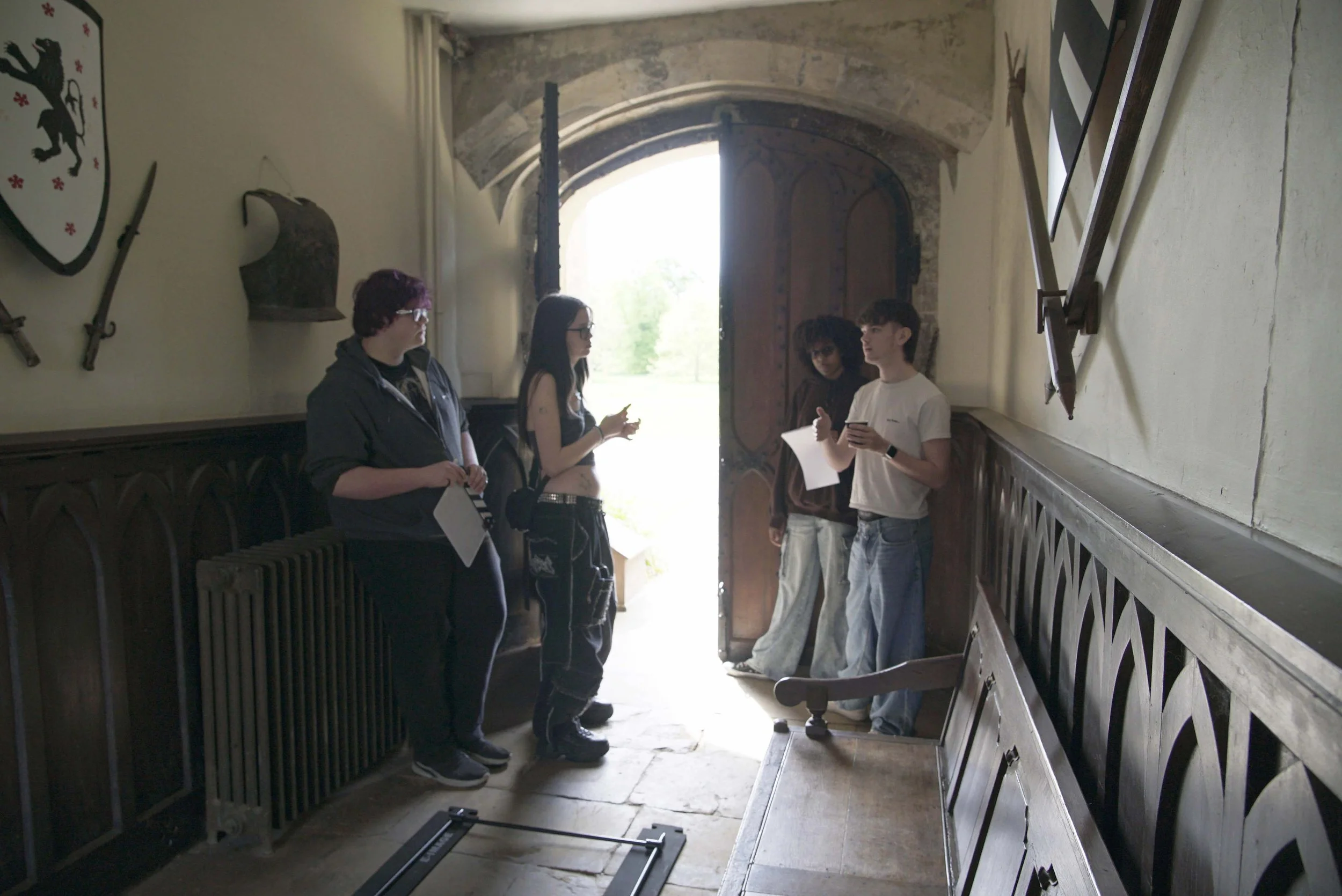 Four young people are standing inside a historic building near an open door with light streaming in. They appear to be engaged in conversation, with one person holding a smartphone. The interior features medieval weaponry and armor displayed on the w