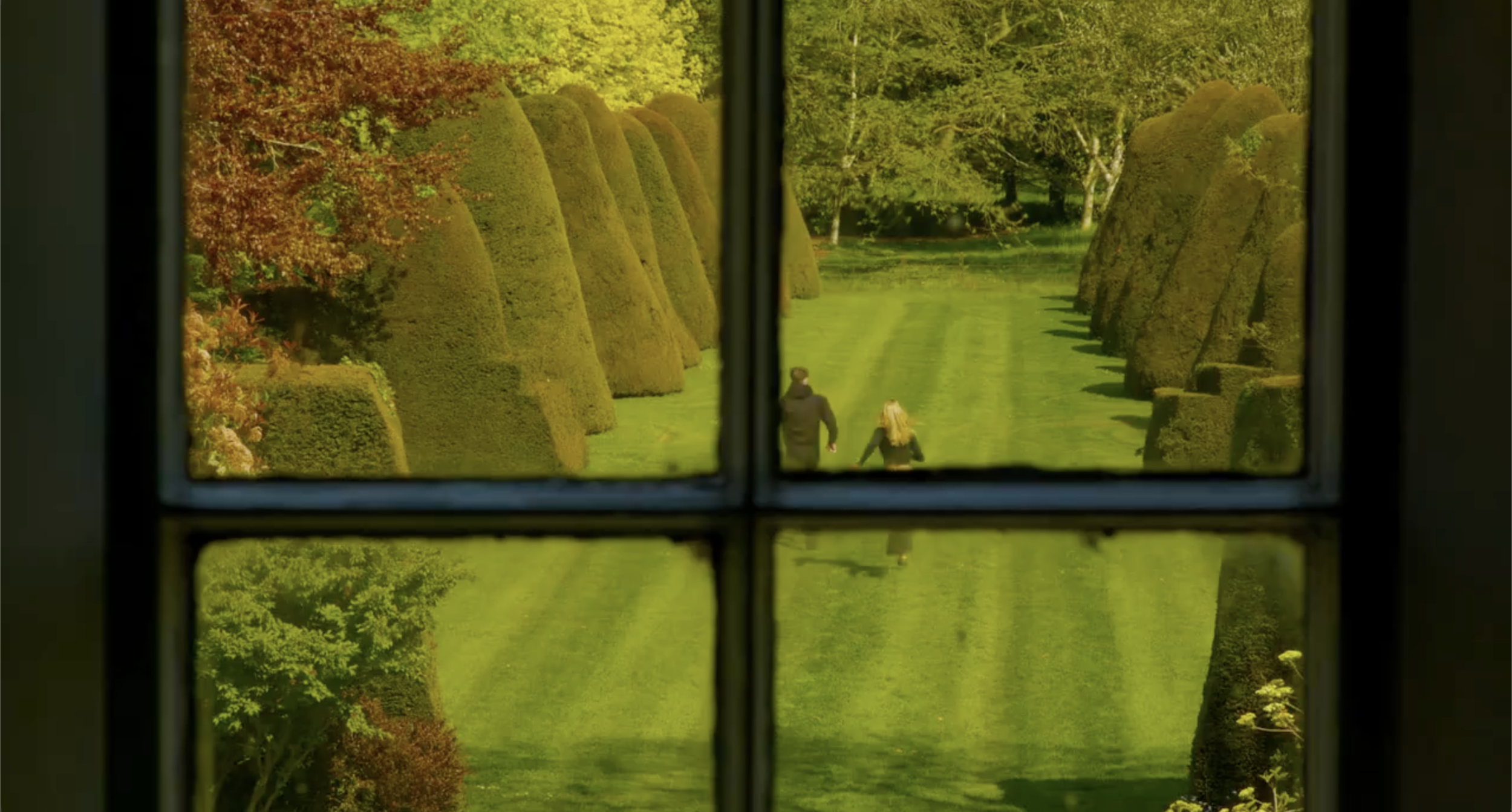 View through window panes showing a garden with trimmed hedges, green grass, and trees, with a couple walking along a path.