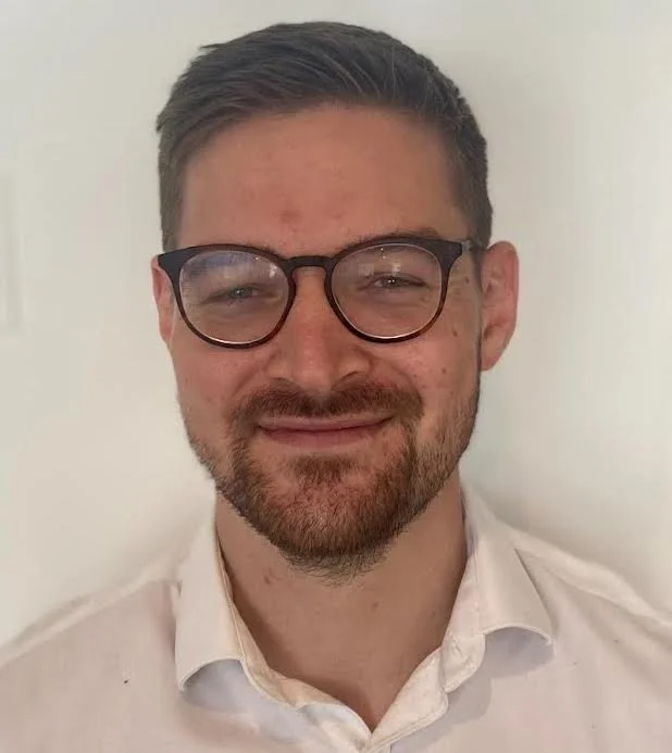 A young man with short brown hair, glasses, and a beard, smiling at the camera, wearing a white collared shirt, against a plain background.
