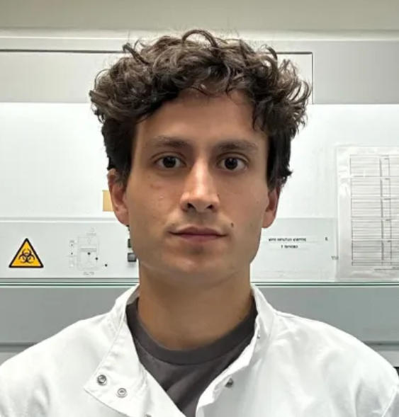 A young man with curly brown hair wearing a white lab coat standing in front of electrical equipment with warning signs.