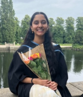 A young woman in a graduation gown holding a bouquet of flowers outdoors near a body of water.